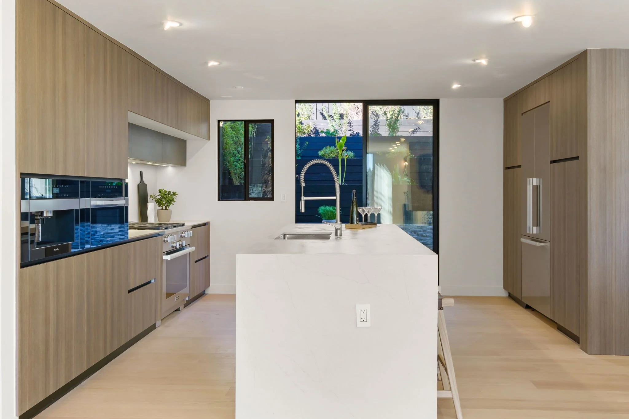 Modern kitchen with wooden cabinets, white countertops, stainless steel appliances, a sink with a tall faucet, and large windows showing outdoor greenery.
