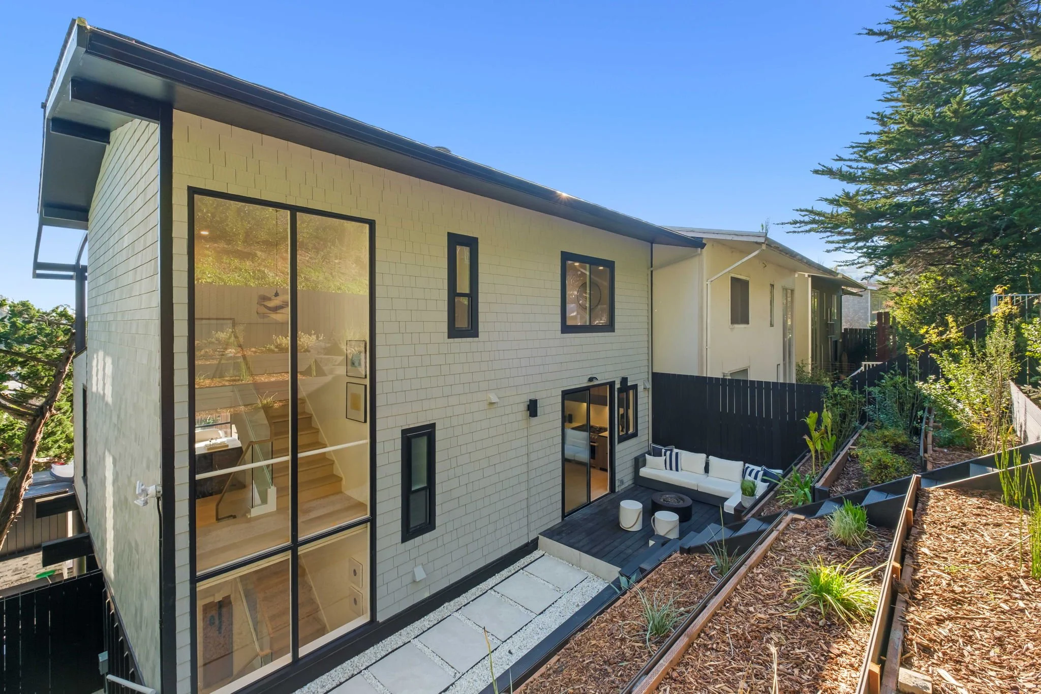 Modern two-story house with a large glass window and sliding door, small outdoor seating area with a white couch and black coffee table on a wooden deck, hillside yard with plants and mulch, bright sunny sky.