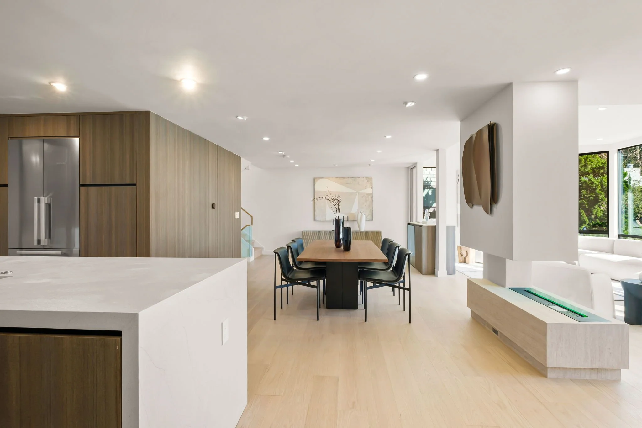 Open-concept modern living and dining area with white walls, light wood flooring, a wooden dining table with black chairs, a fireplace, large windows, and minimalistic artwork.