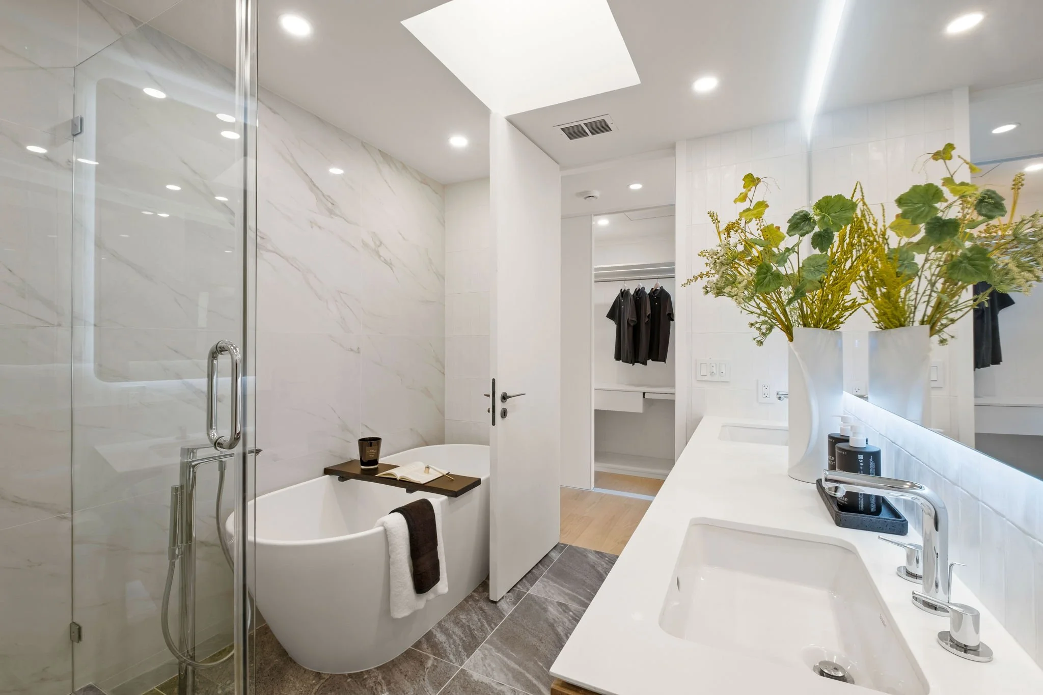 Modern bathroom with white marble walls, a walk-in shower, a freestanding bathtub, a countertop with a sink, and a large vase with green plants.