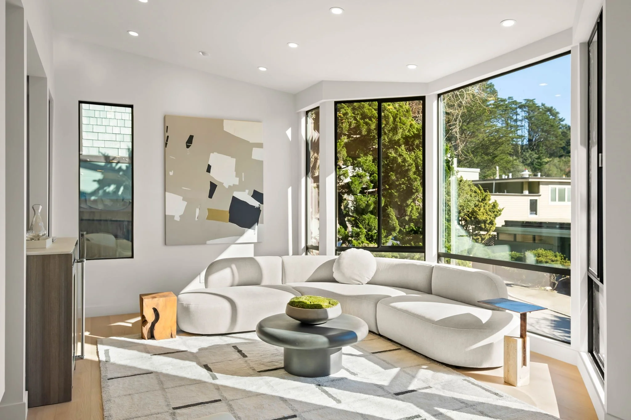 Bright living room with large windows, white curved sofa, abstract wall art, wooden side table, and a modern coffee table with a bowl of moss, sunlight streaming in.