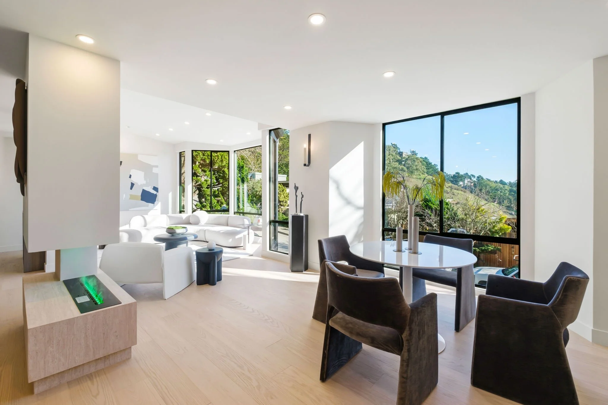 Modern living room with large windows, white sofa, black dining chairs, round white table, and decorative vases with greenery.