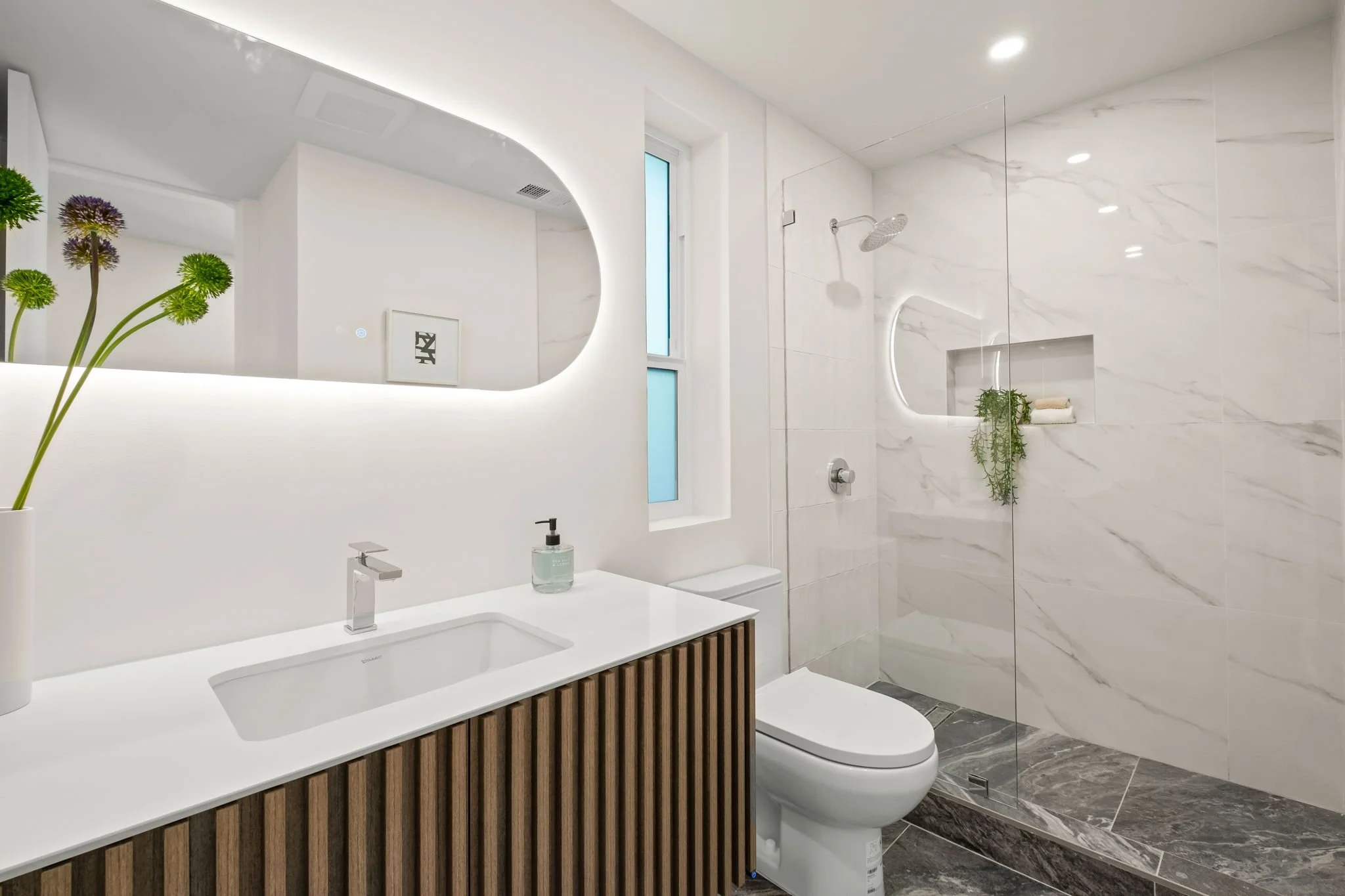 Modern white bathroom with wooden vanity, large mirror, toilet, shower with glass partition, built-in shelf, and minimal decor.