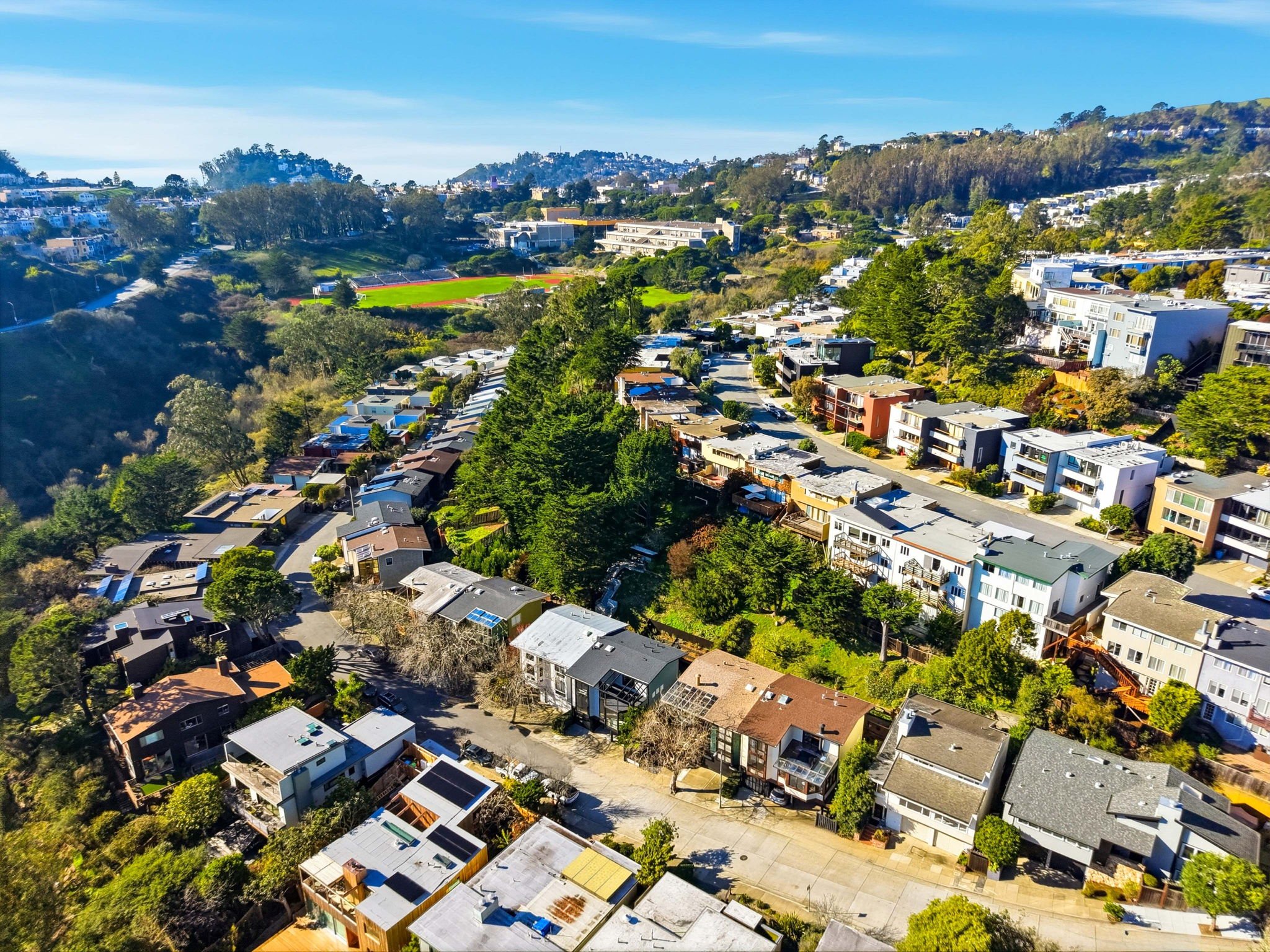 Aerial view of a neighborhood with houses, trees, and a sports field in the distance under a blue sky.