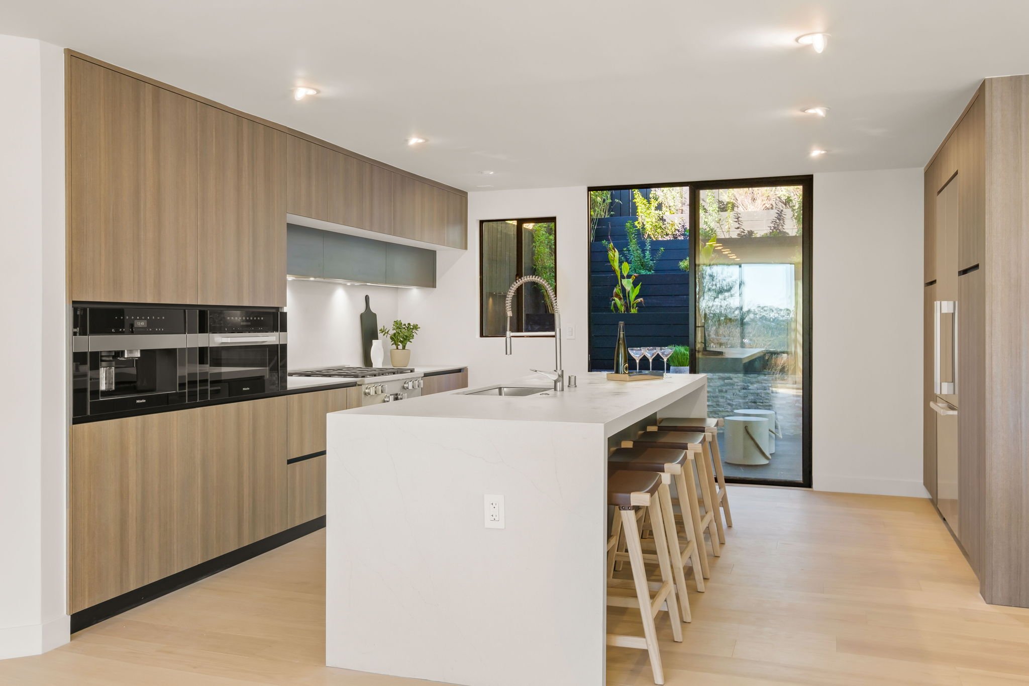 Modern kitchen with wooden cabinets, white marble island, black appliances, and large windows overlooking a patio with greenery.