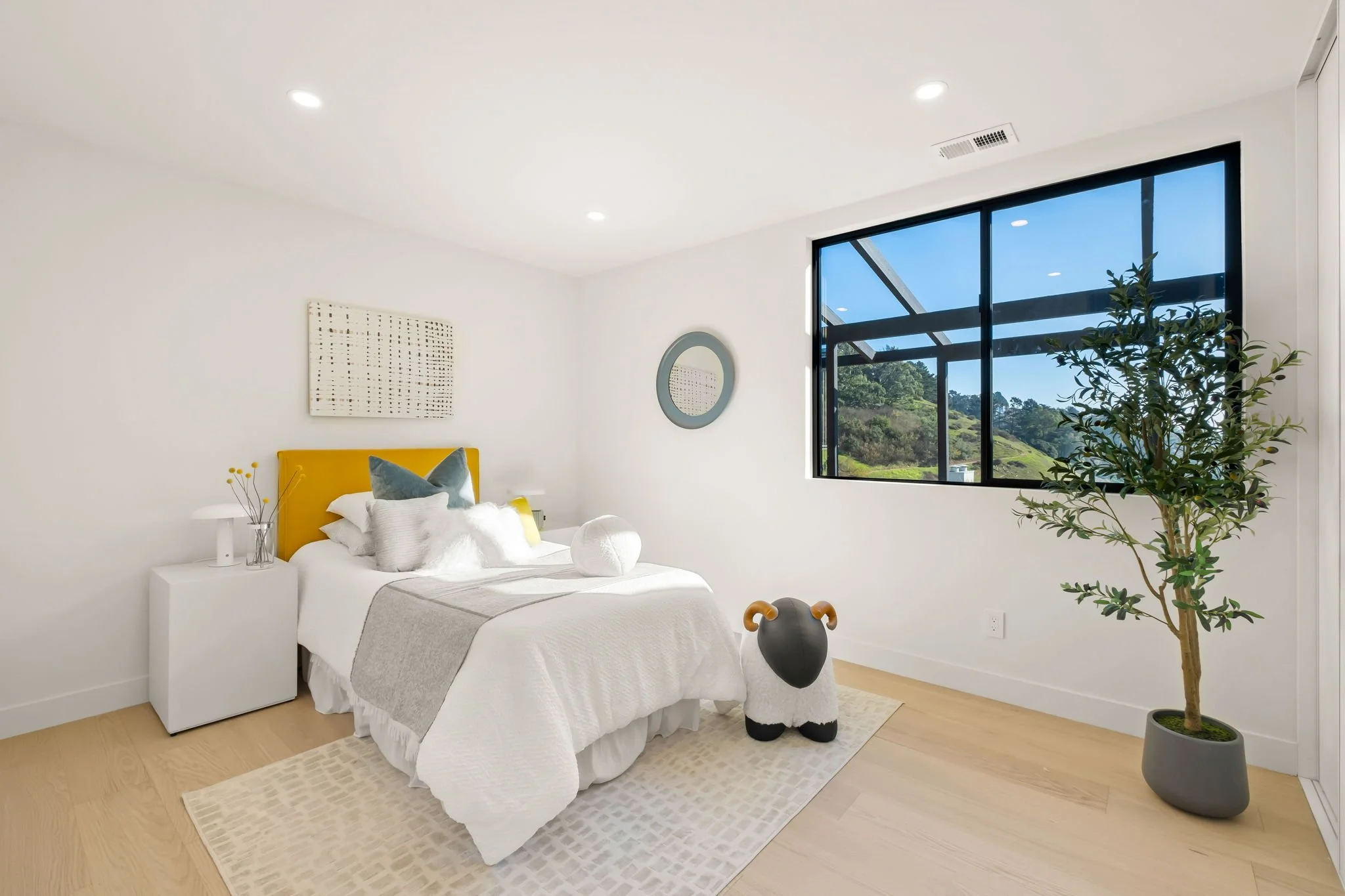 Bright bedroom with white walls, yellow headboard, large window showing greenery, bed with white bedding and pillows, a small white side table with yellow flowers, a sheep-shaped pouf, a potted plant, and wall art.