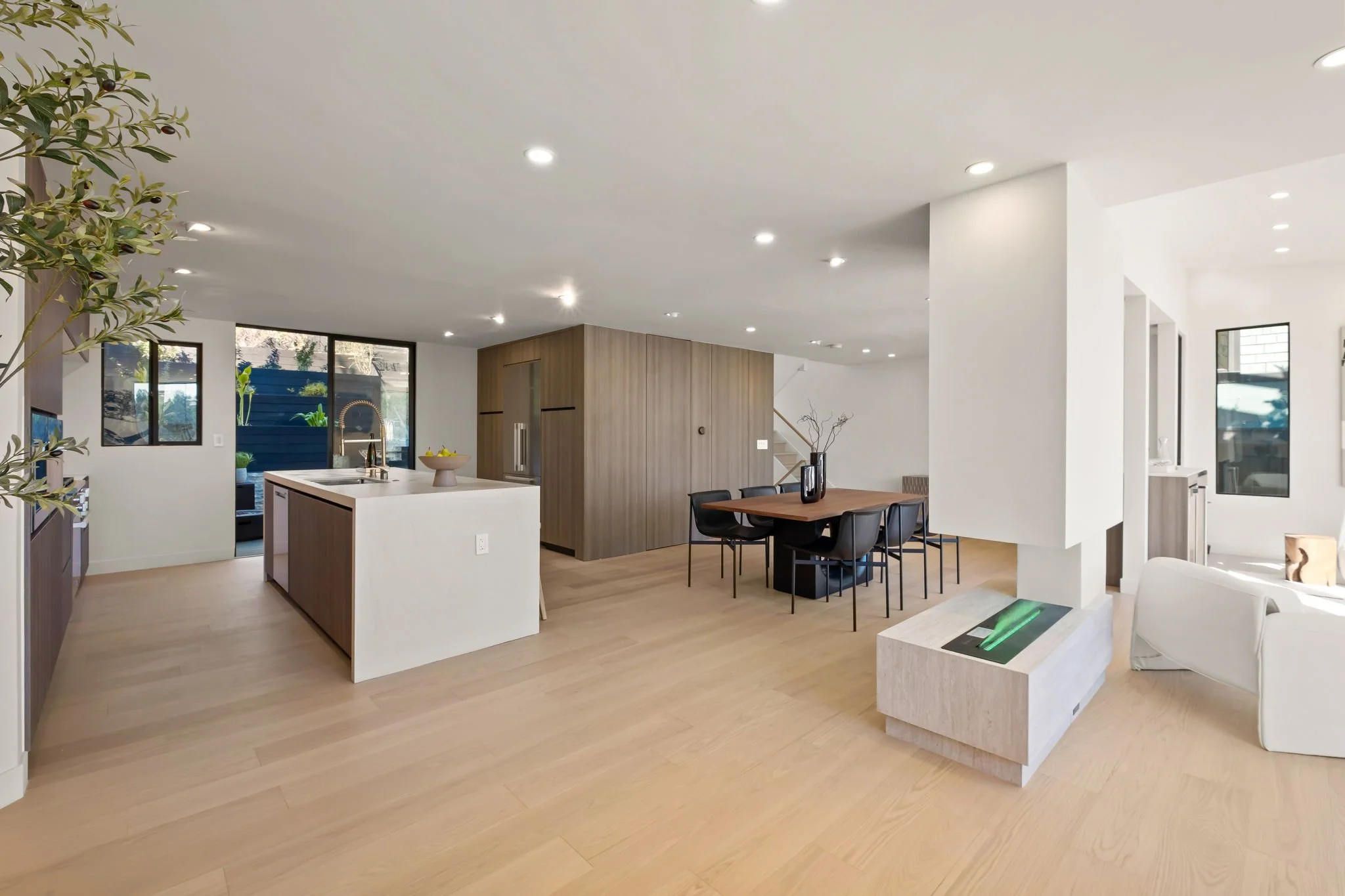 Open-concept modern living room and kitchen with light wood floors, white walls, and large windows; kitchen features an island with a sink, and a white countertop; dining area has a wooden table with black chairs; decorative vases and plant in the sp