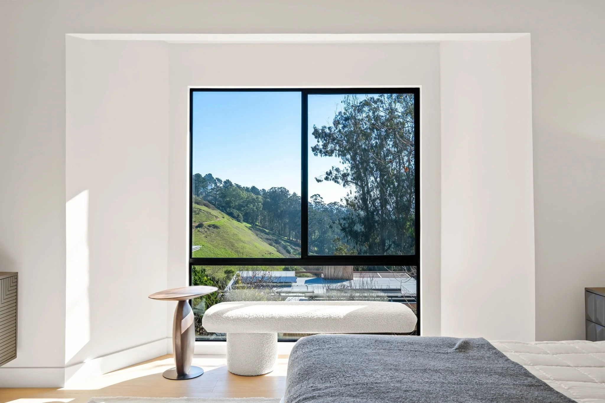 Modern bedroom with large window showing green hillside and trees, white bench, small wooden side table, bed with gray blanket, minimal decor.