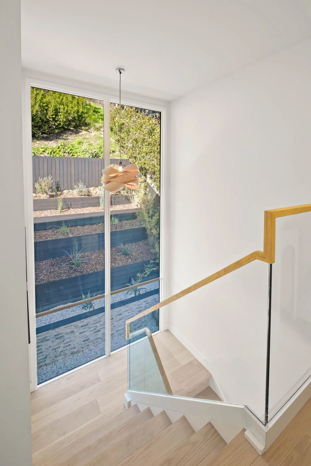 Interior view of a staircase with wooden handrail, white walls, and a large window overlooking a garden with plants and shrubs.