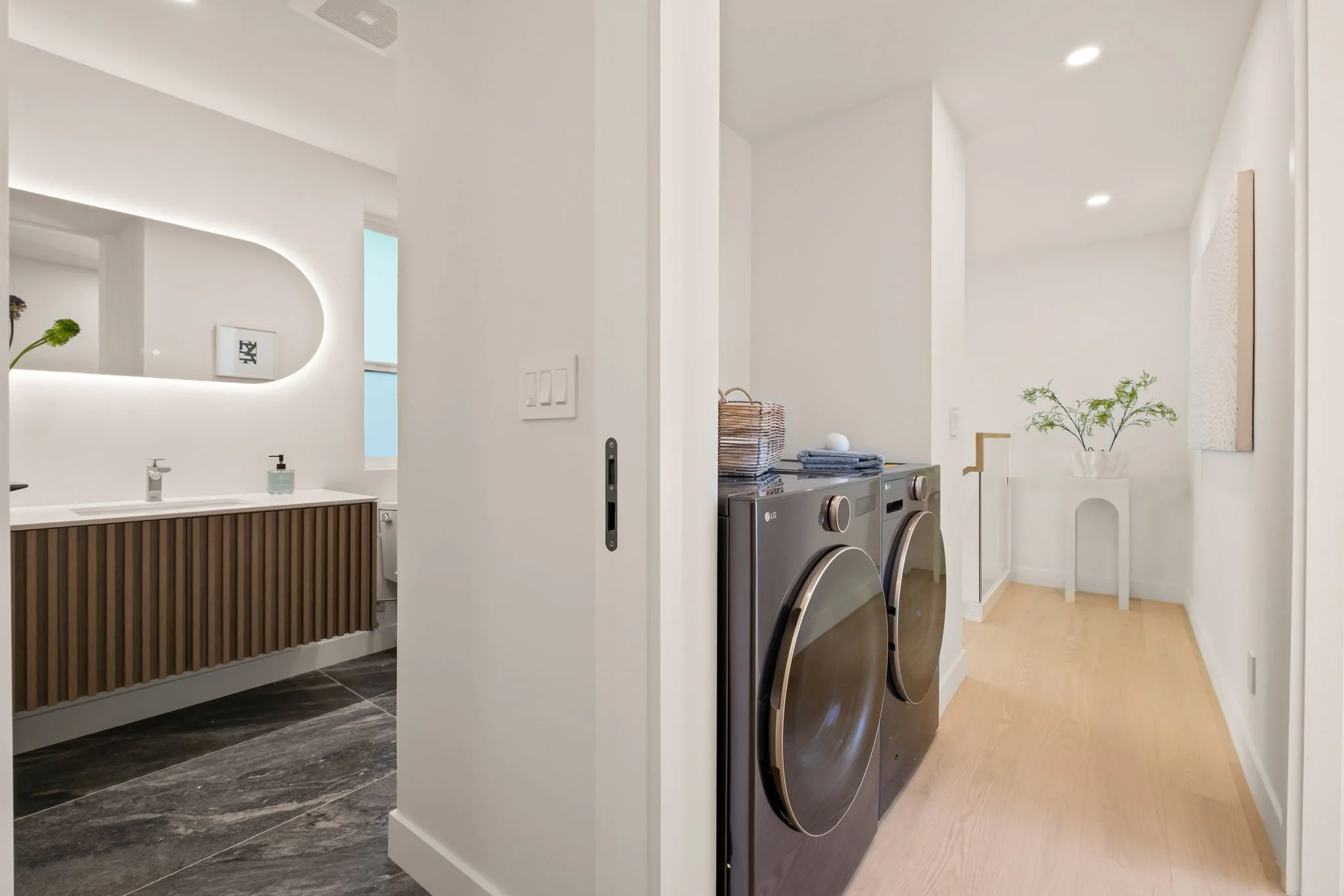 A laundry area with a front-loading washer and dryer, stacked on top of each other, on a light wooden floor. There's a small window with a plant and sculpture on a white stand, and minimal decor with white walls.