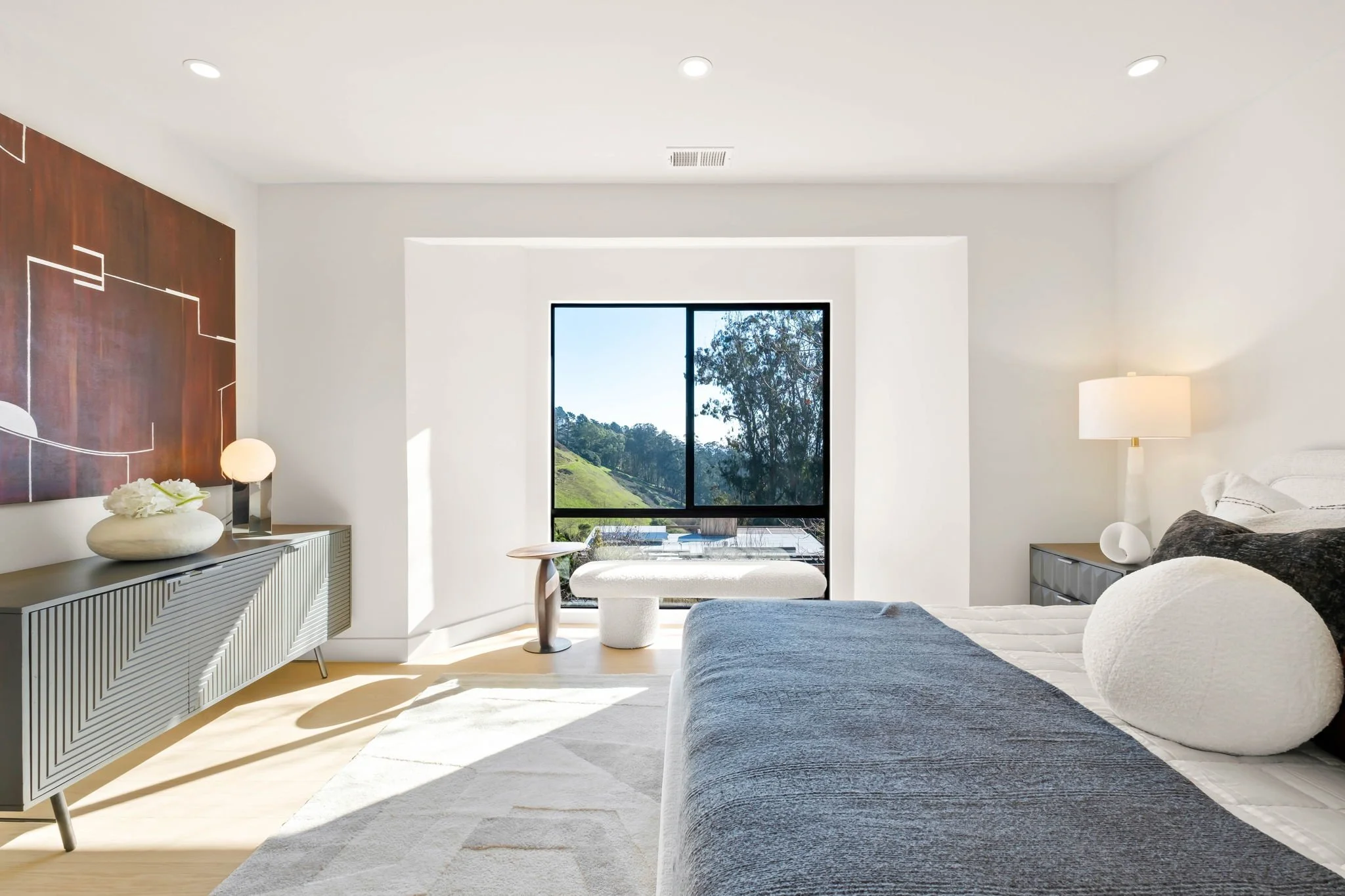 Modern bedroom with white walls, large window showing outdoor greenery, bed with white and dark gray bedding, nightstand with white lamp, dresser with decorative objects, and abstract artwork on the wall.