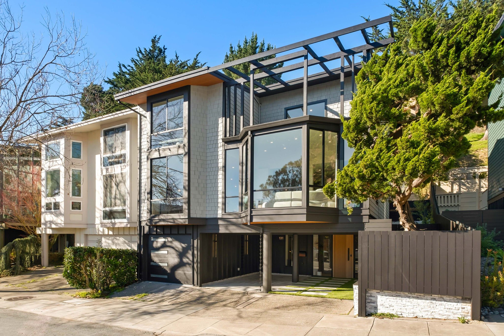 Modern multi-story house with large glass windows, white and black exterior, and a partially covered driveway. A tree and shrubs are in front, with a blue sky above.