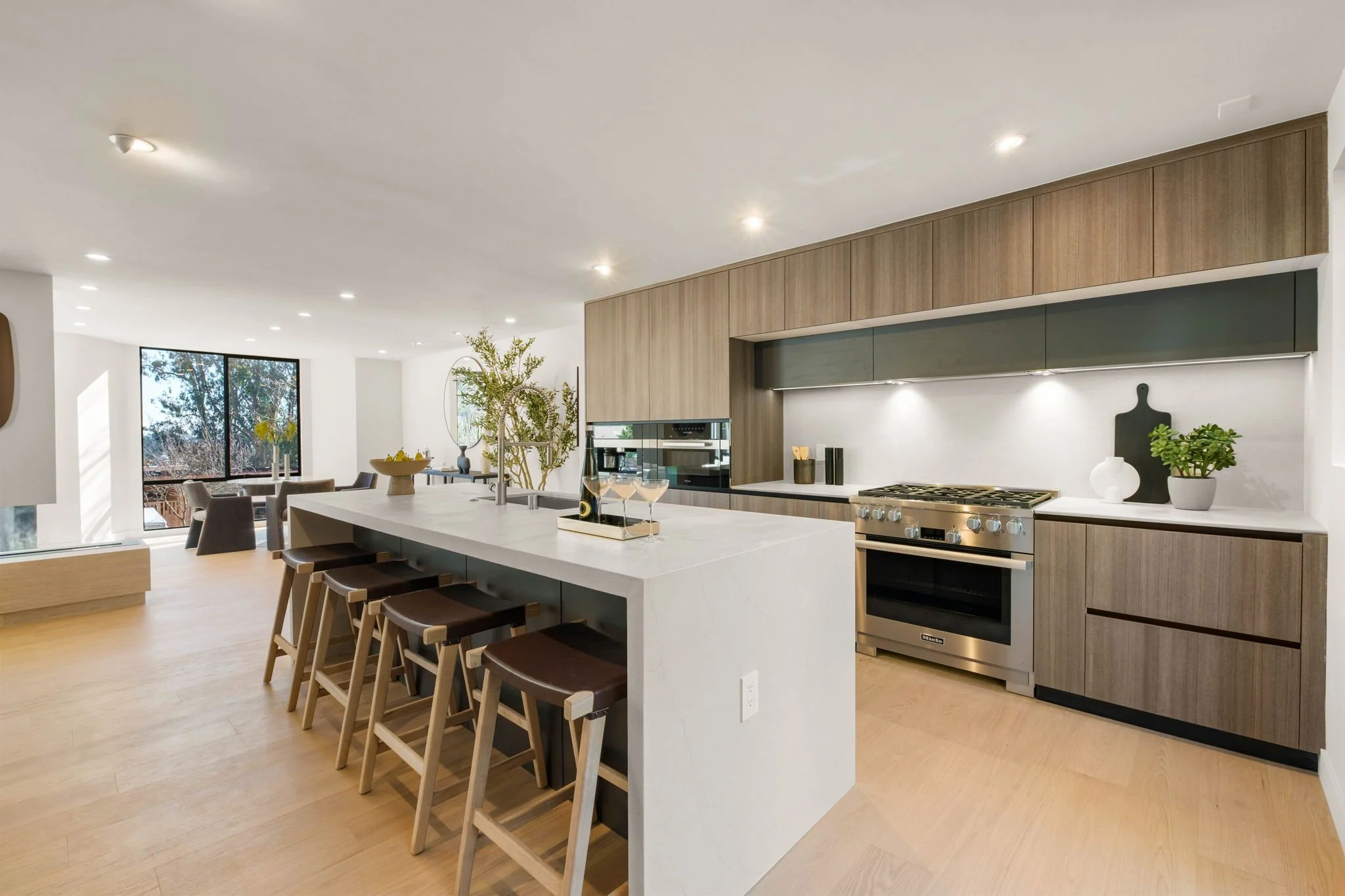 Modern open-concept kitchen with wood cabinets, stainless steel appliances, white island with bar stools, and a dining area near large windows in a bright, minimalist home.