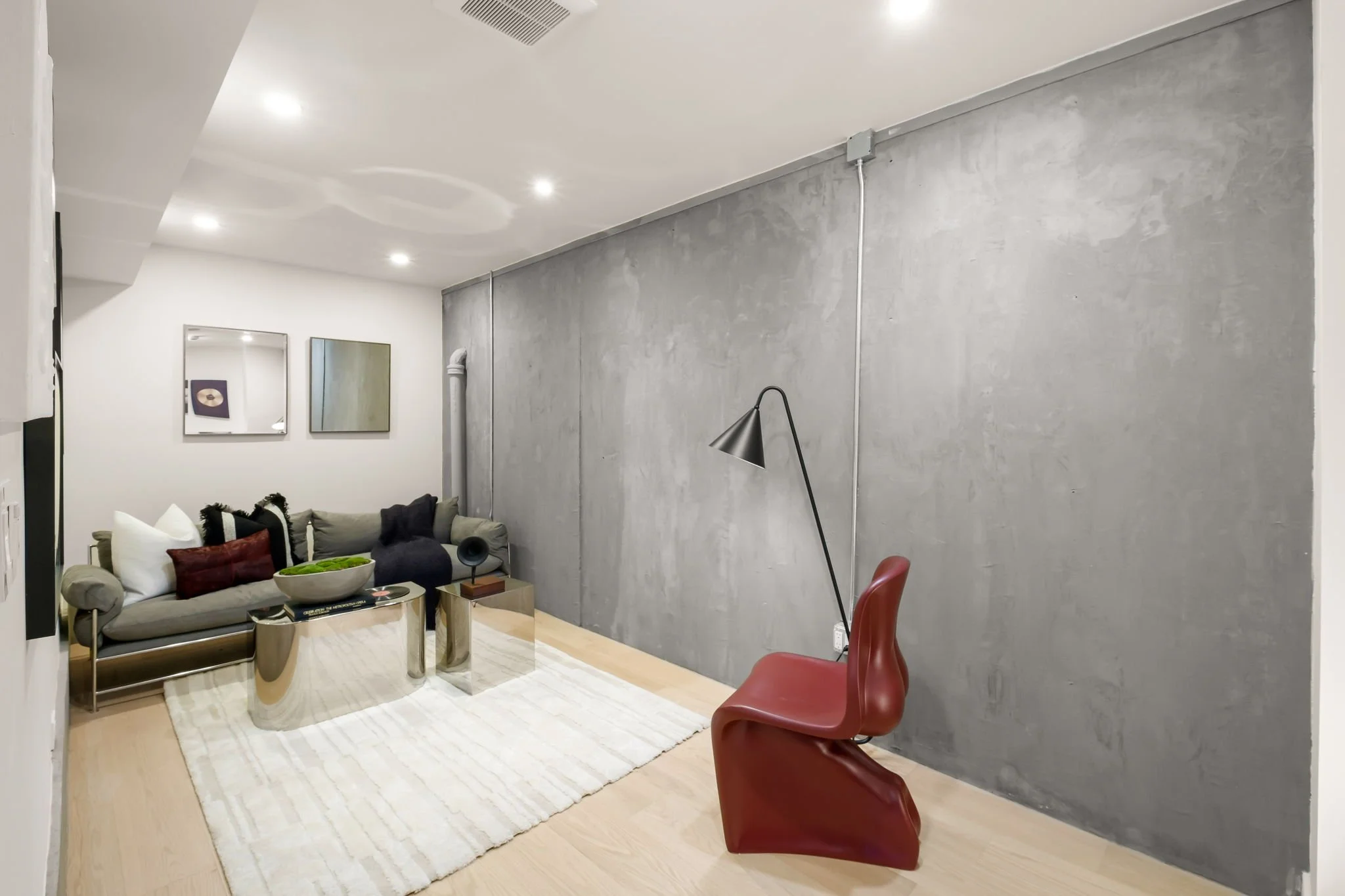 Modern living room with gray sofa, black and white cushions, a red chair, a glass coffee table, a white rug, and minimalist decor with framed artwork on a white wall, and a concrete accent wall.