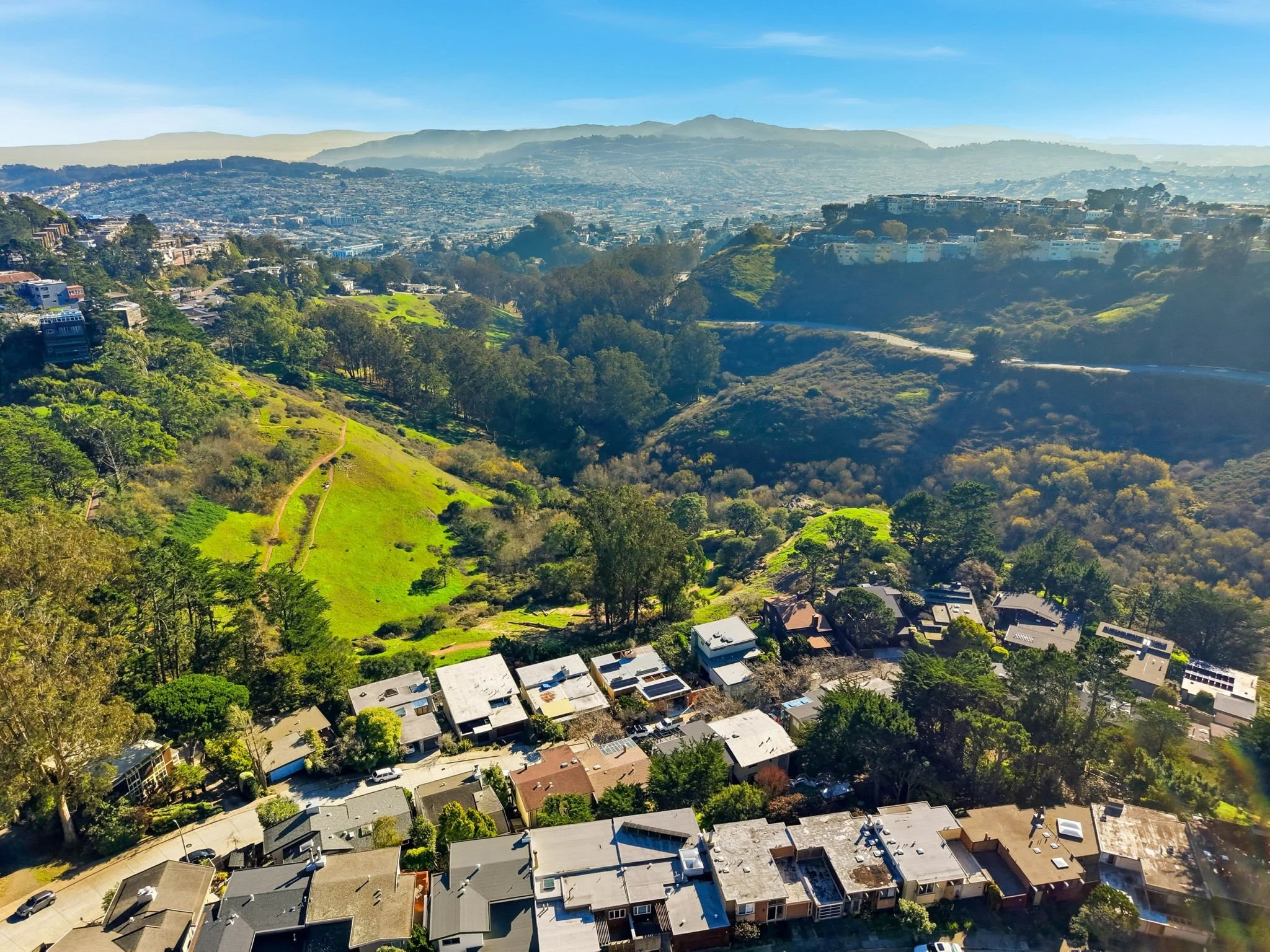 Aerial view of a hilly neighborhood with houses, green trees, and a large grassy park surrounded by hills and distant cityscape under a clear blue sky.
