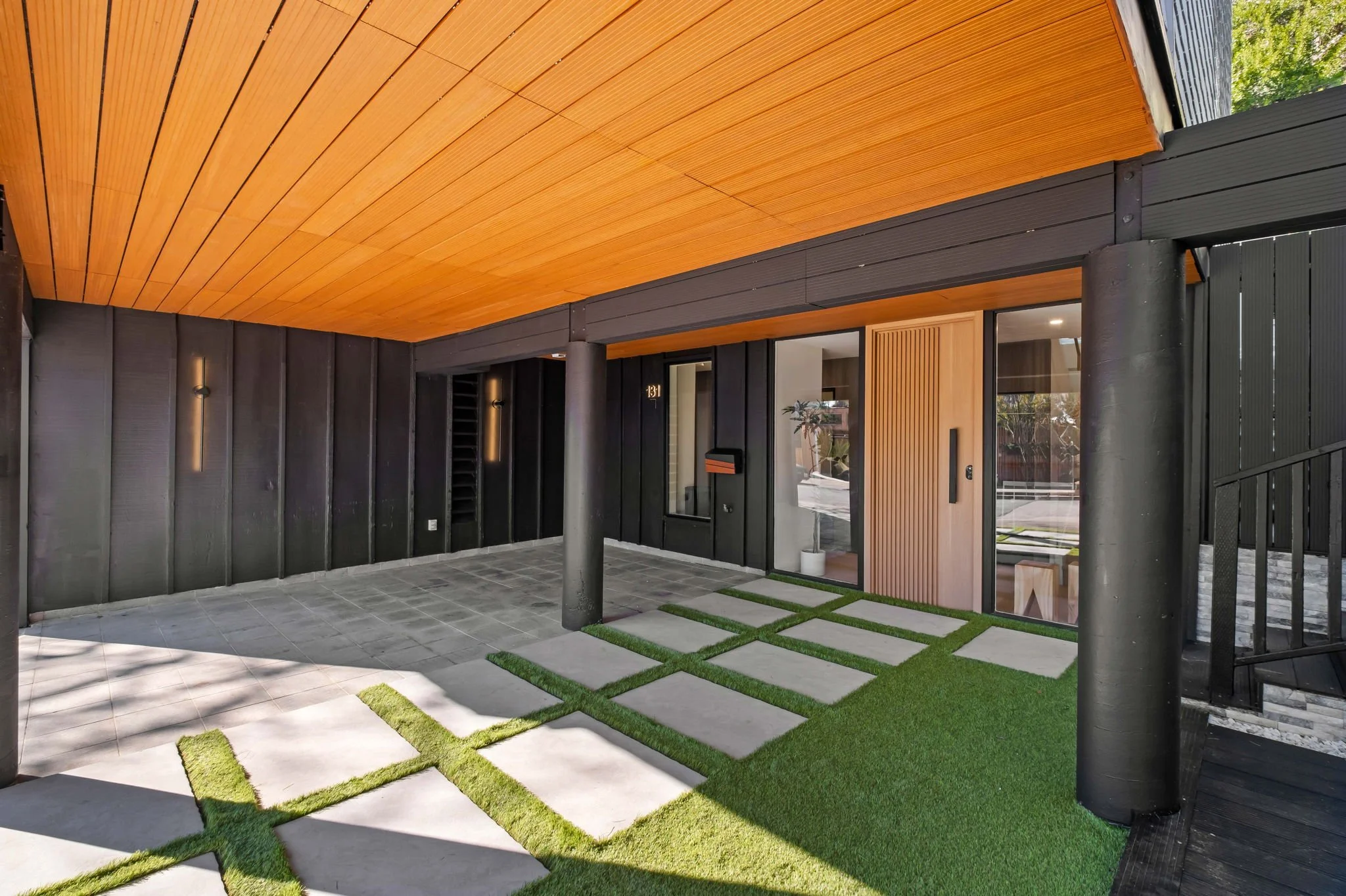 Modern house entrance with black paneled walls, large glass door, a wooden accent, and a small lawn with concrete stepping stones.