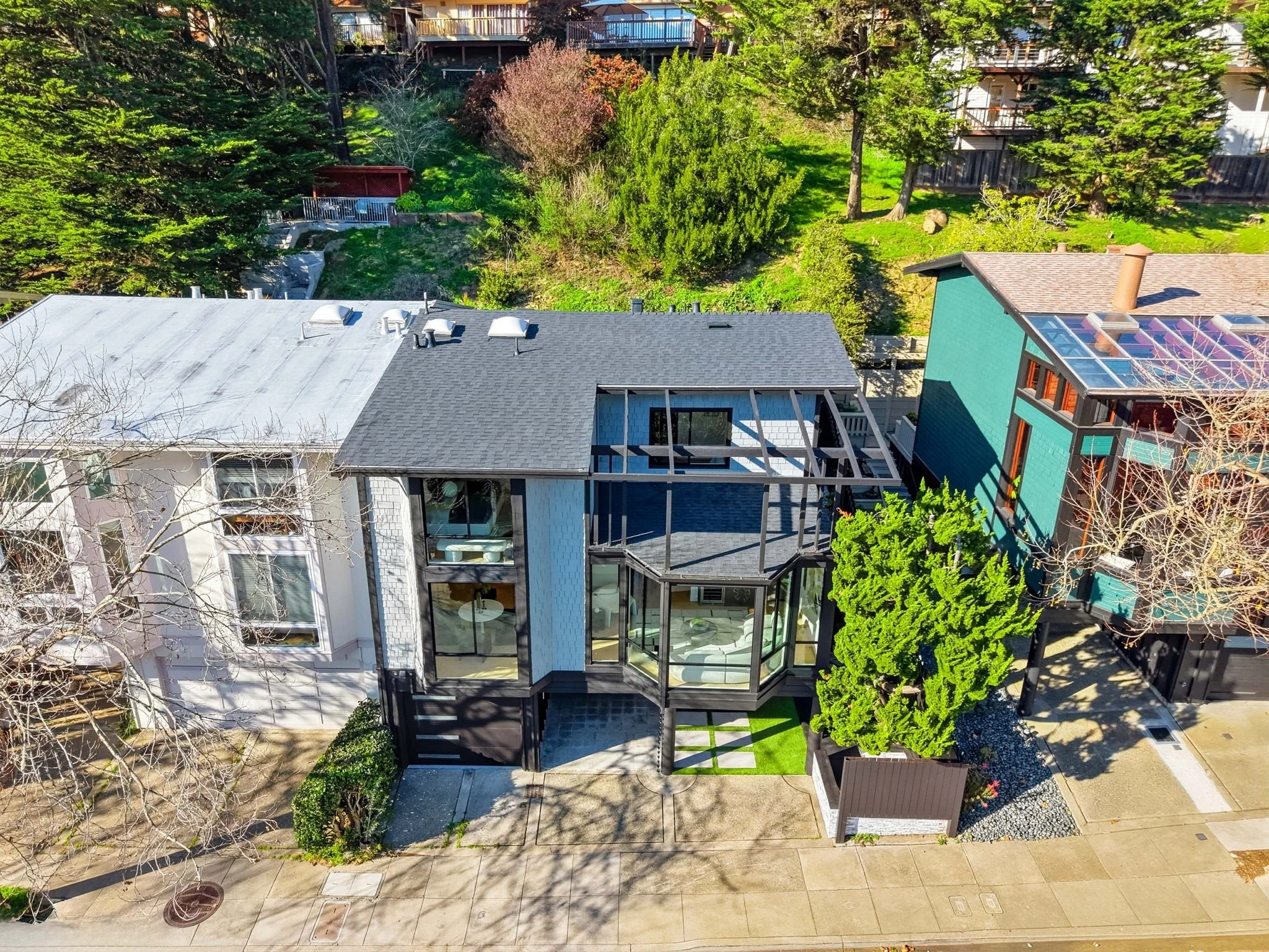 Aerial view of a modern two-story house with large glass windows, surrounded by trees and neighboring houses, with a sidewalk in front.