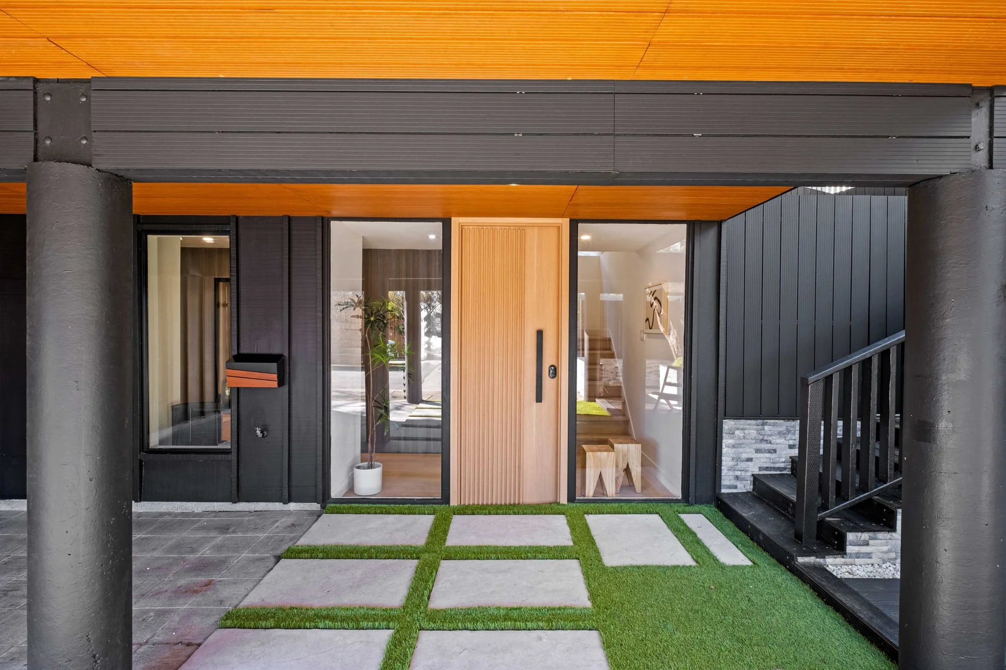 Modern house entryway with glass door, stone pathway, and small patches of artificial grass, black exterior walls, and wood accents.