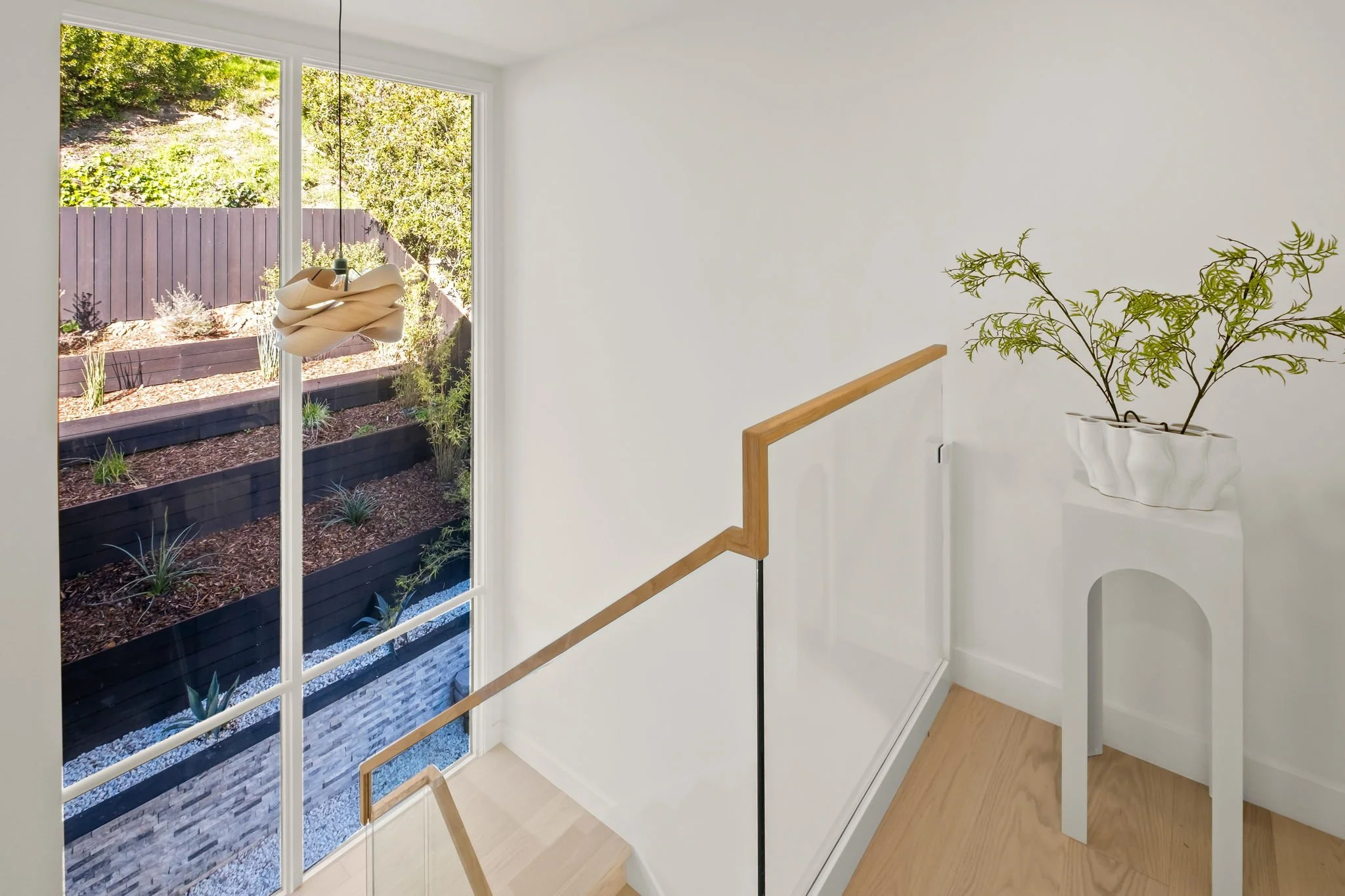 Interior view of a staircase landing with a wooden handrail and a large window overlooking a garden with tiered black raised beds, plants, and mulch. A decorative white planter with a green plant is on a white pedestal.