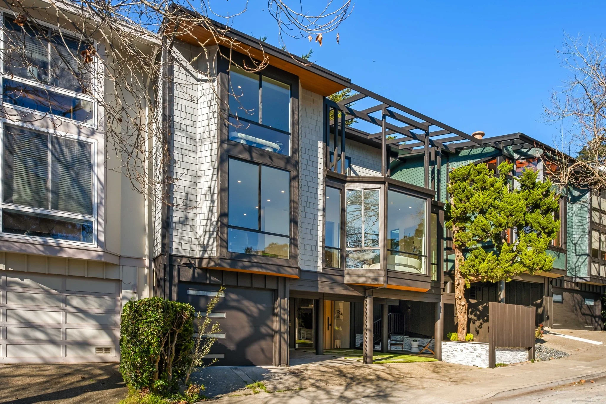 Modern multi-story residential building with large glass windows and a small front yard, under a clear blue sky.