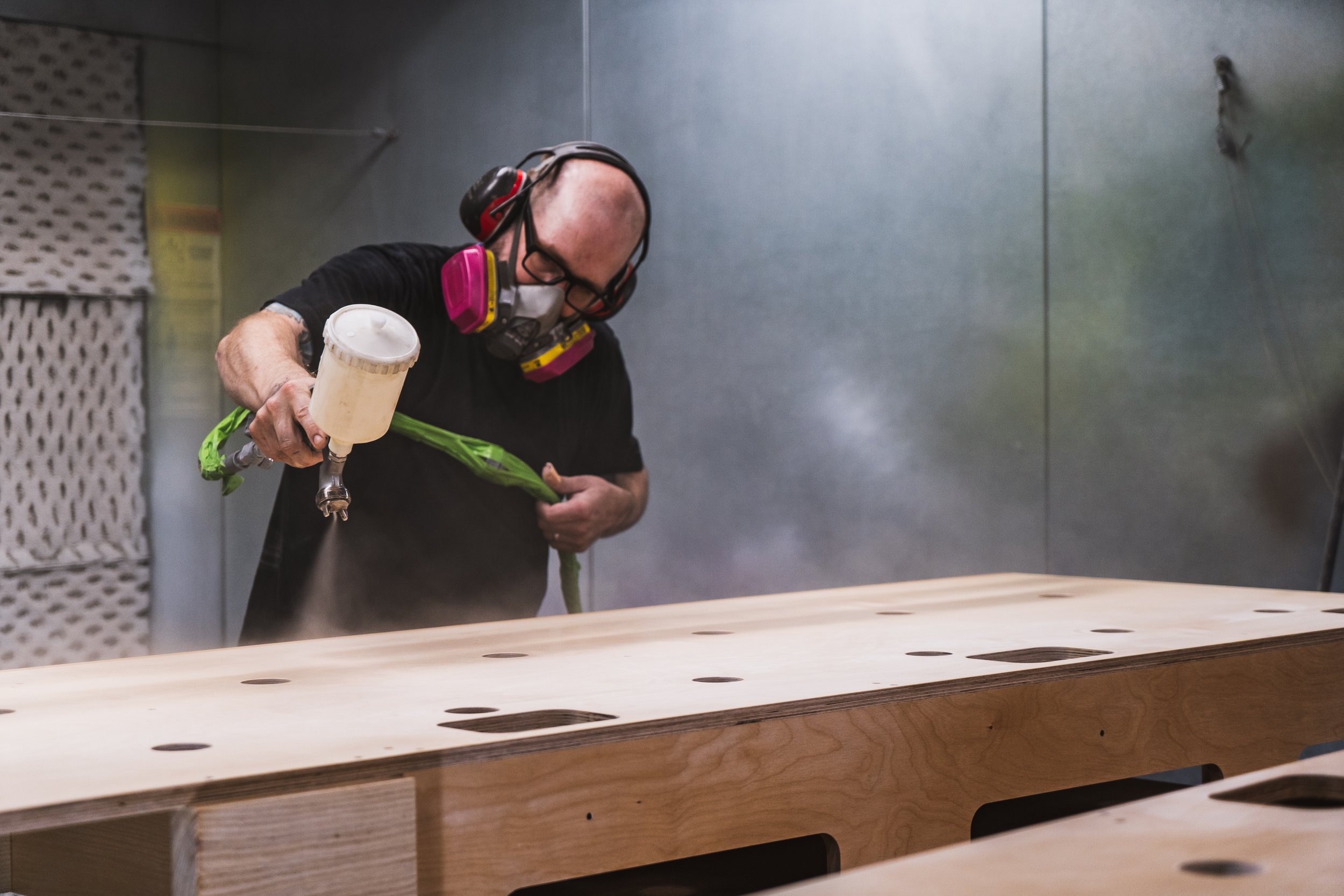 A man wearing a respirator mask and glasses is spray-painting a wooden piece in a workshop.