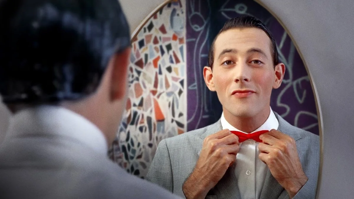 A man in a suit is tying a red bow tie while looking at his reflection in a mirror.