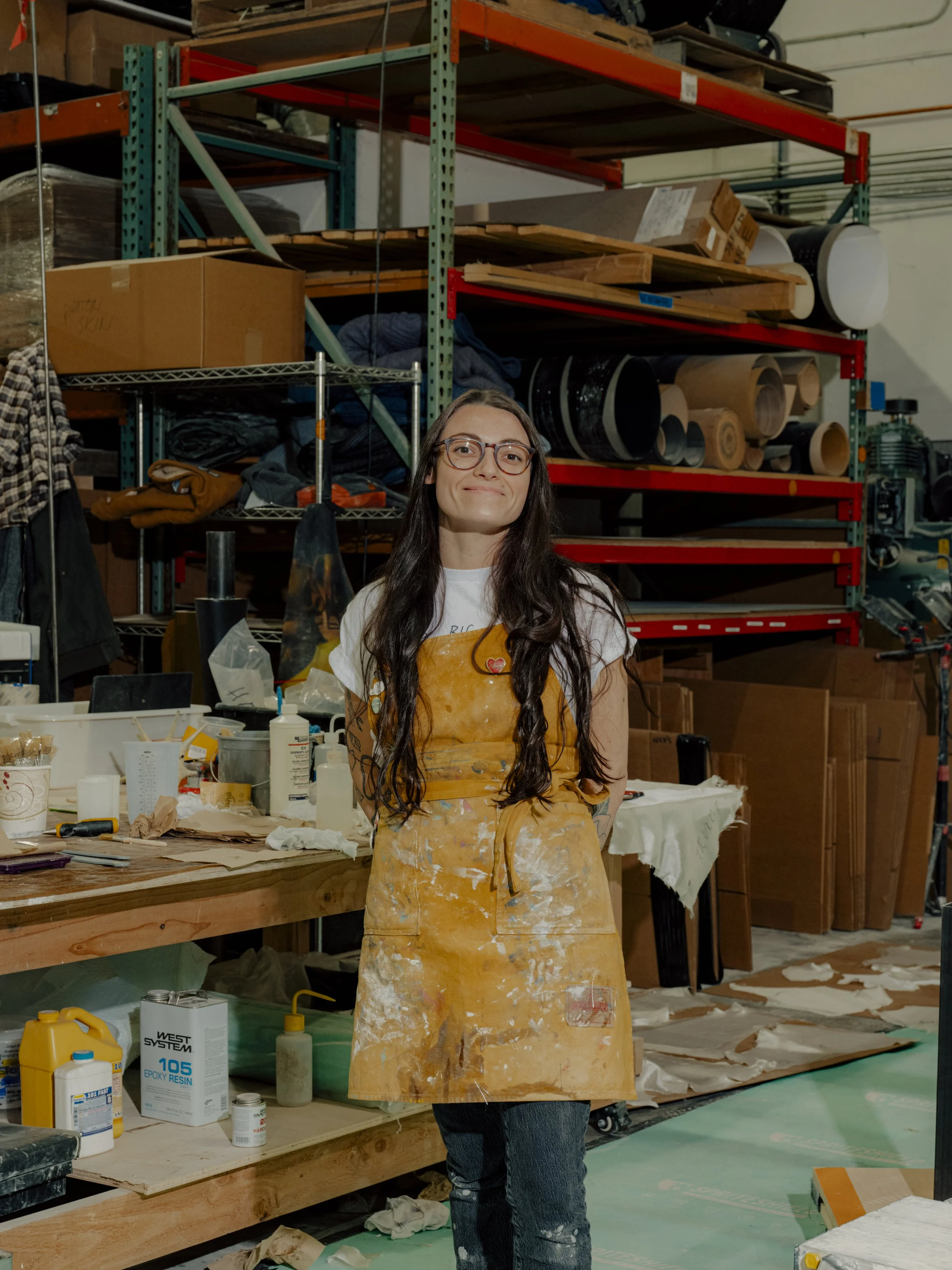 A woman with long dark hair and glasses standing in a workshop or studio, wearing a worn yellow apron covered in white paint and dirt, with tools and materials on a workbench behind her, and industrial shelving with various supplies in the background.