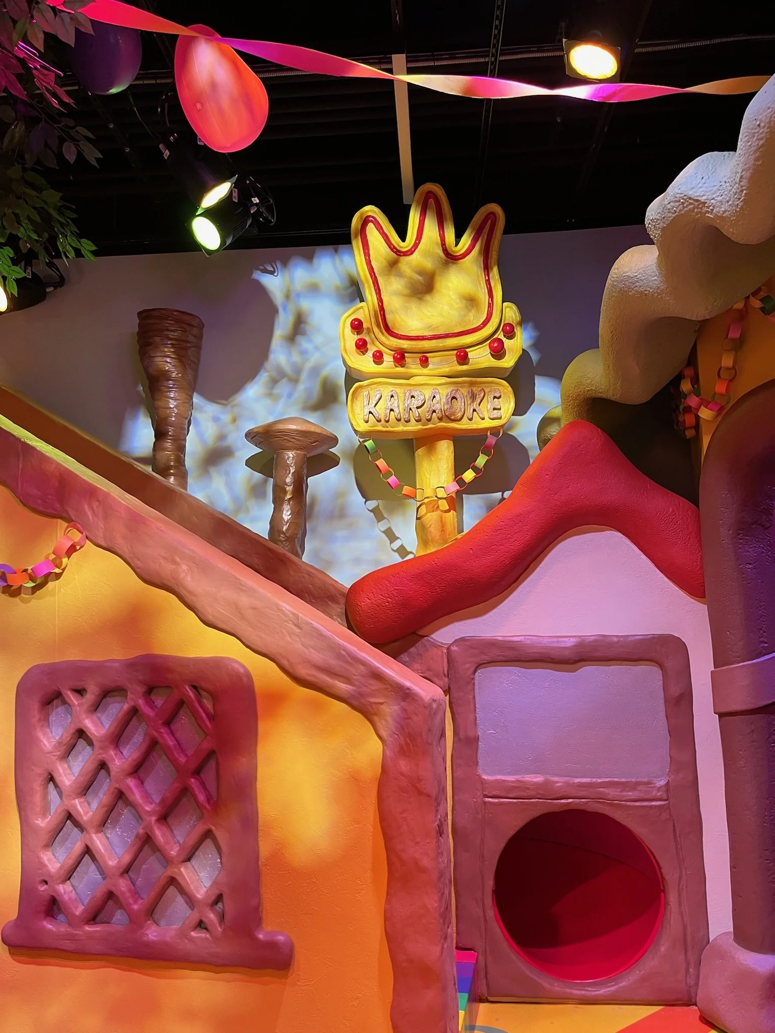 Decorative indoor scene with a yellow illuminated karaoke sign resembling a crown, colorful paper streamers, pink, yellow, and orange abstract shapes, and various textured wall and furniture structures.