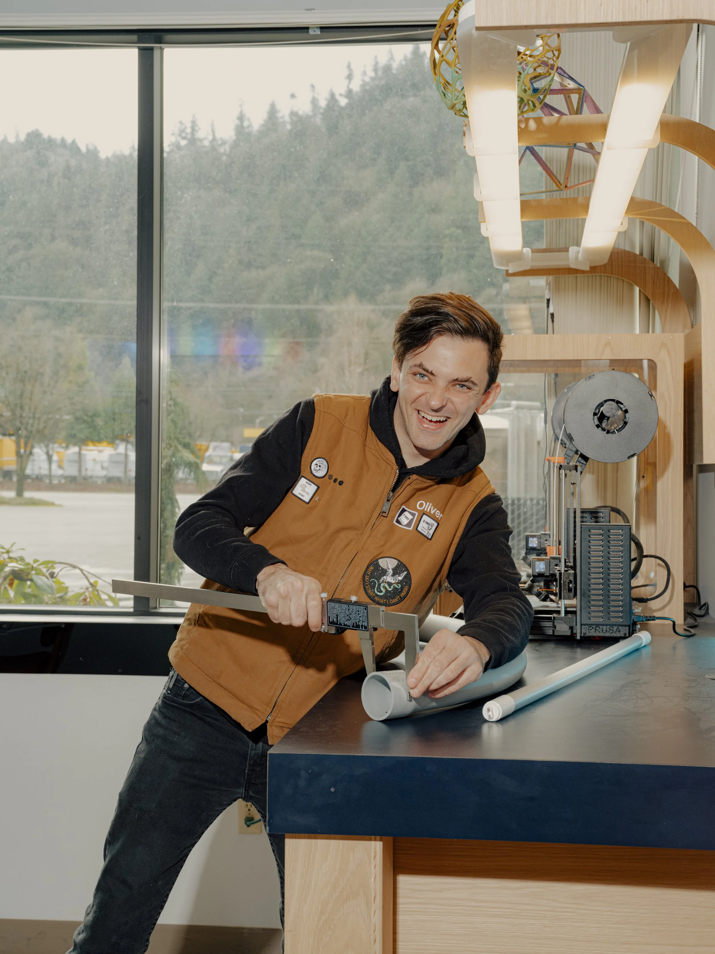 A smiling man working on a science or engineering project at a workshop, using a saw on a long tube of material, with equipment and tools around him and a large window behind.