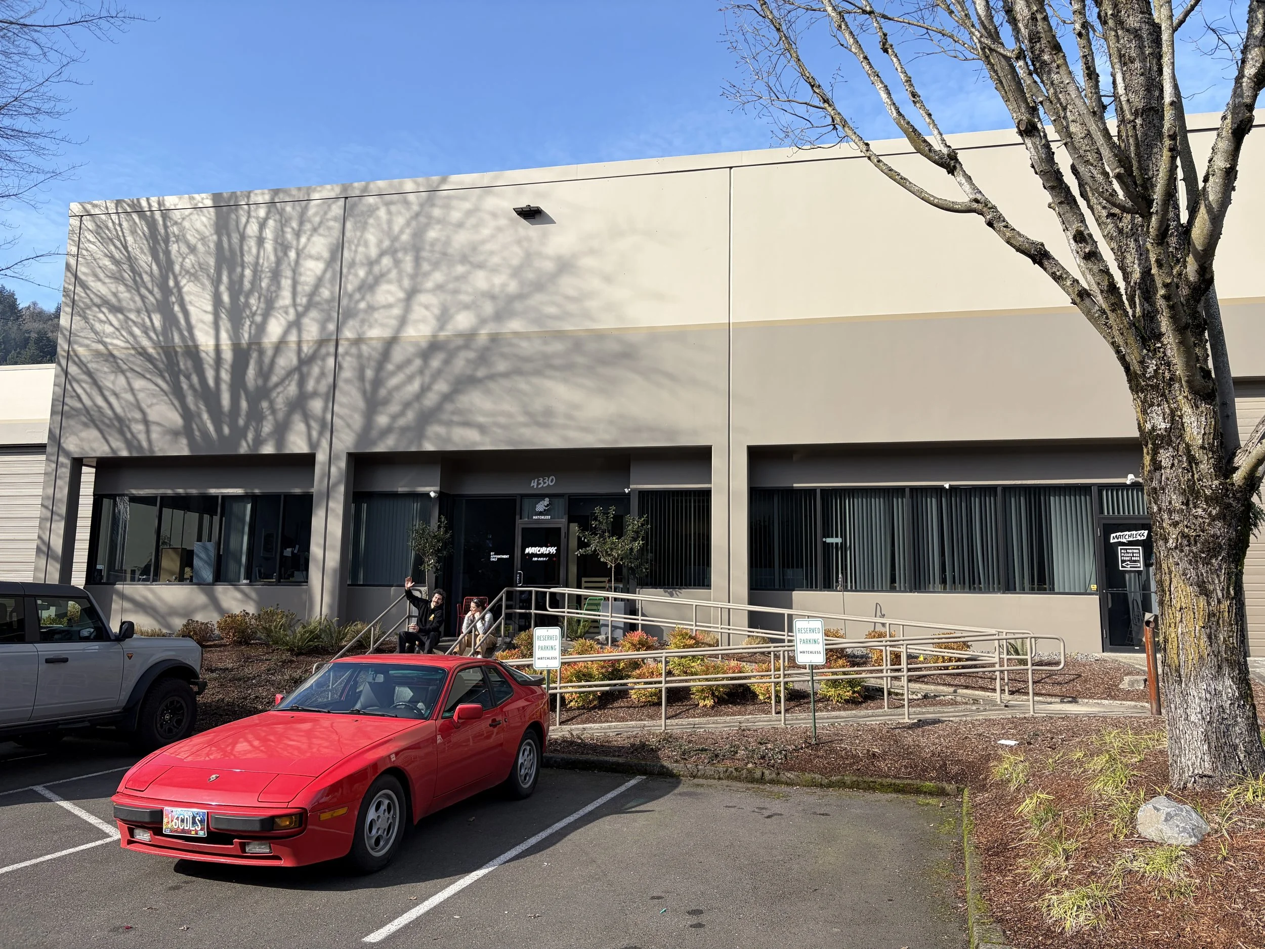 A beige commercial building with large windows, outside a parking lot with several cars including a red sports car. There are two people sitting on the steps near the entrance, one waving. There are parking signs and a tree casting shadows on the building.
