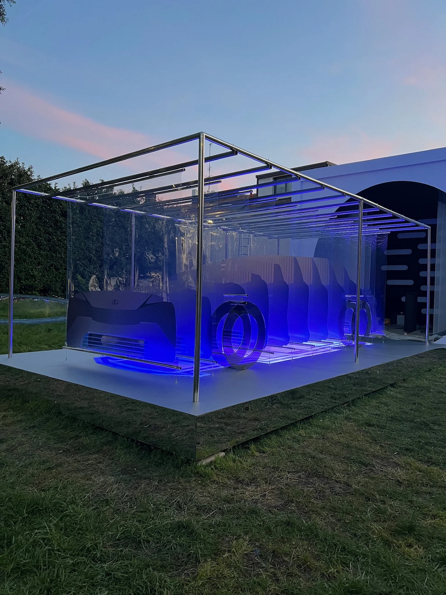 An outdoor display featuring a car-like structure illuminated with blue neon lights, situated on a metallic platform with grass surrounding it.