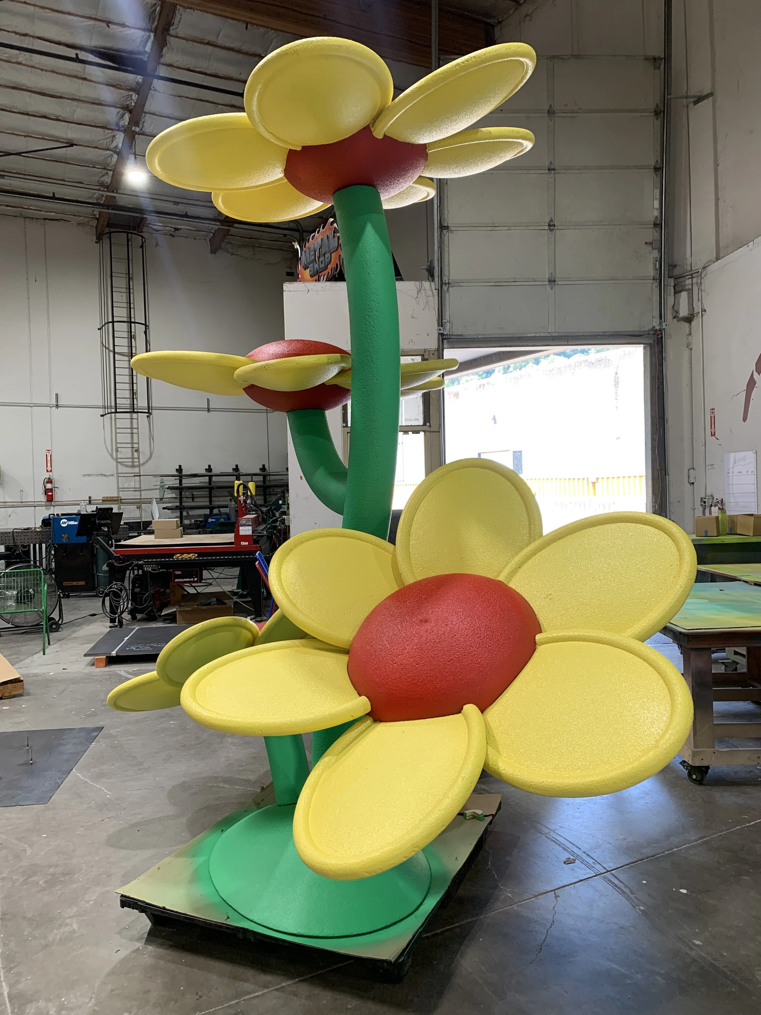 Colorful flower sculpture with yellow petals and red centers, green stems, located inside a warehouse or workshop.