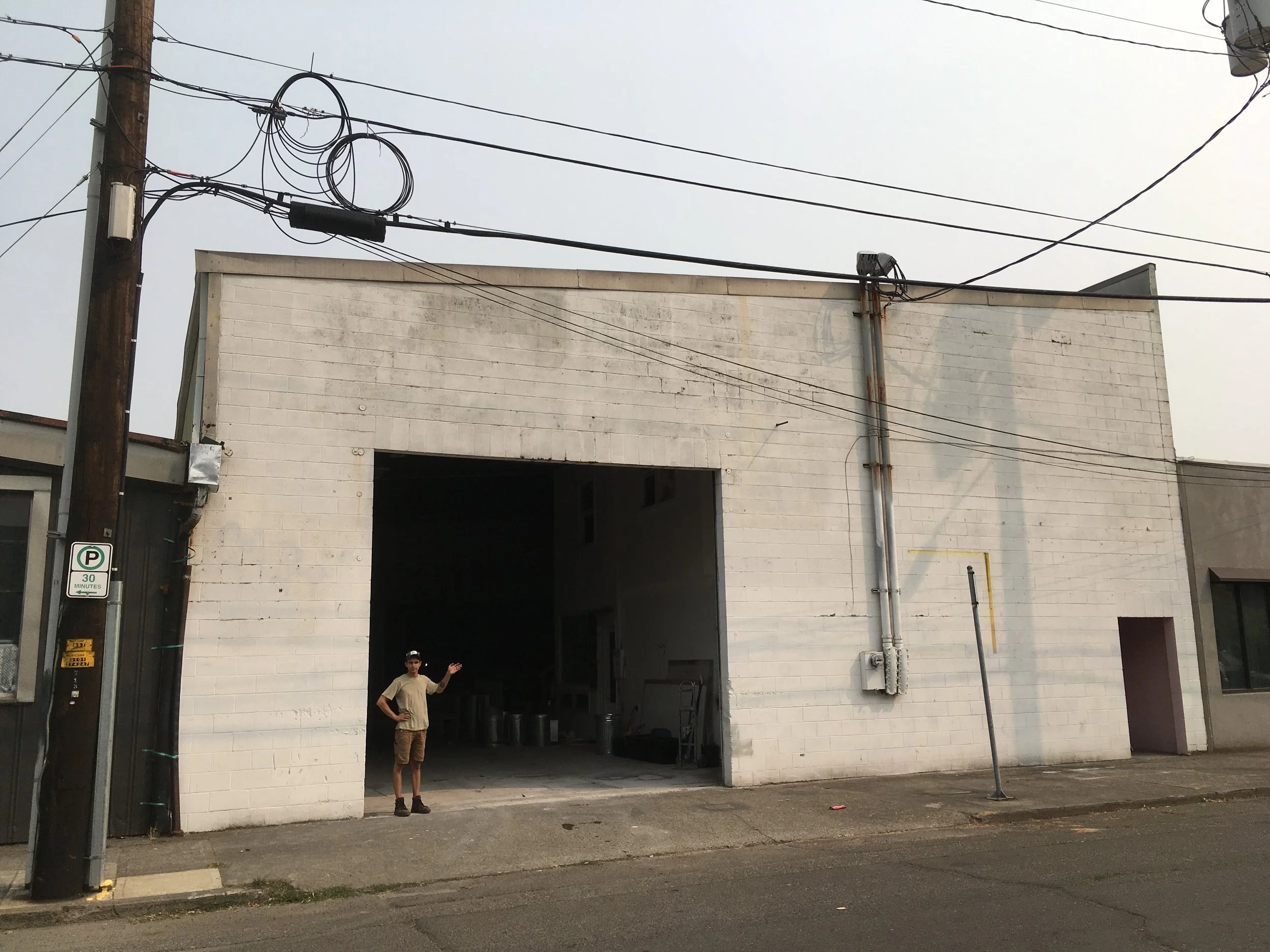 A person standing outside a large white brick building with an open garage door, in an urban area with power lines overhead, and a parking sign on a utility pole.