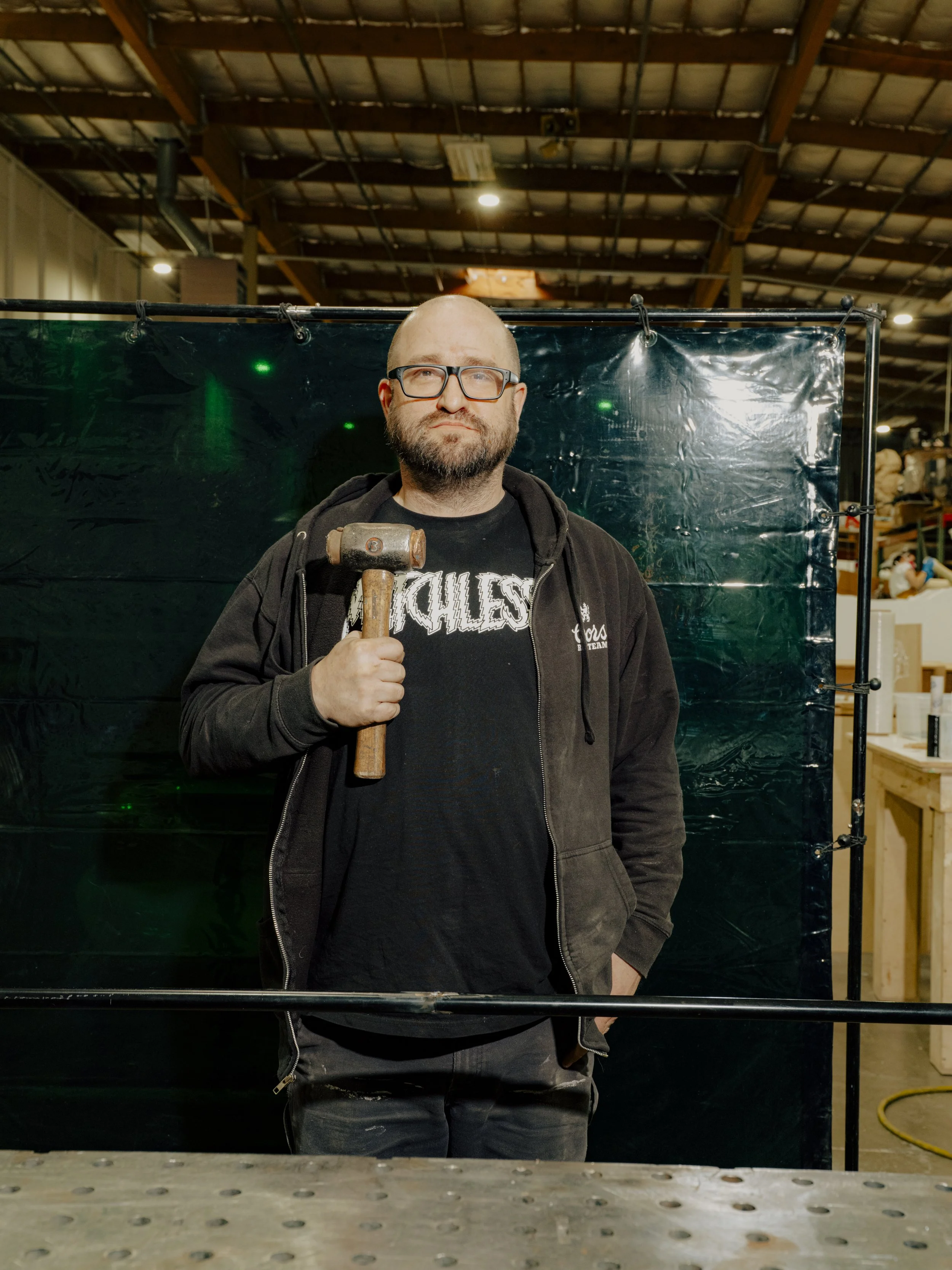 A man with glasses, a beard, and a black hoodie holding a hammer stands in an industrial warehouse.