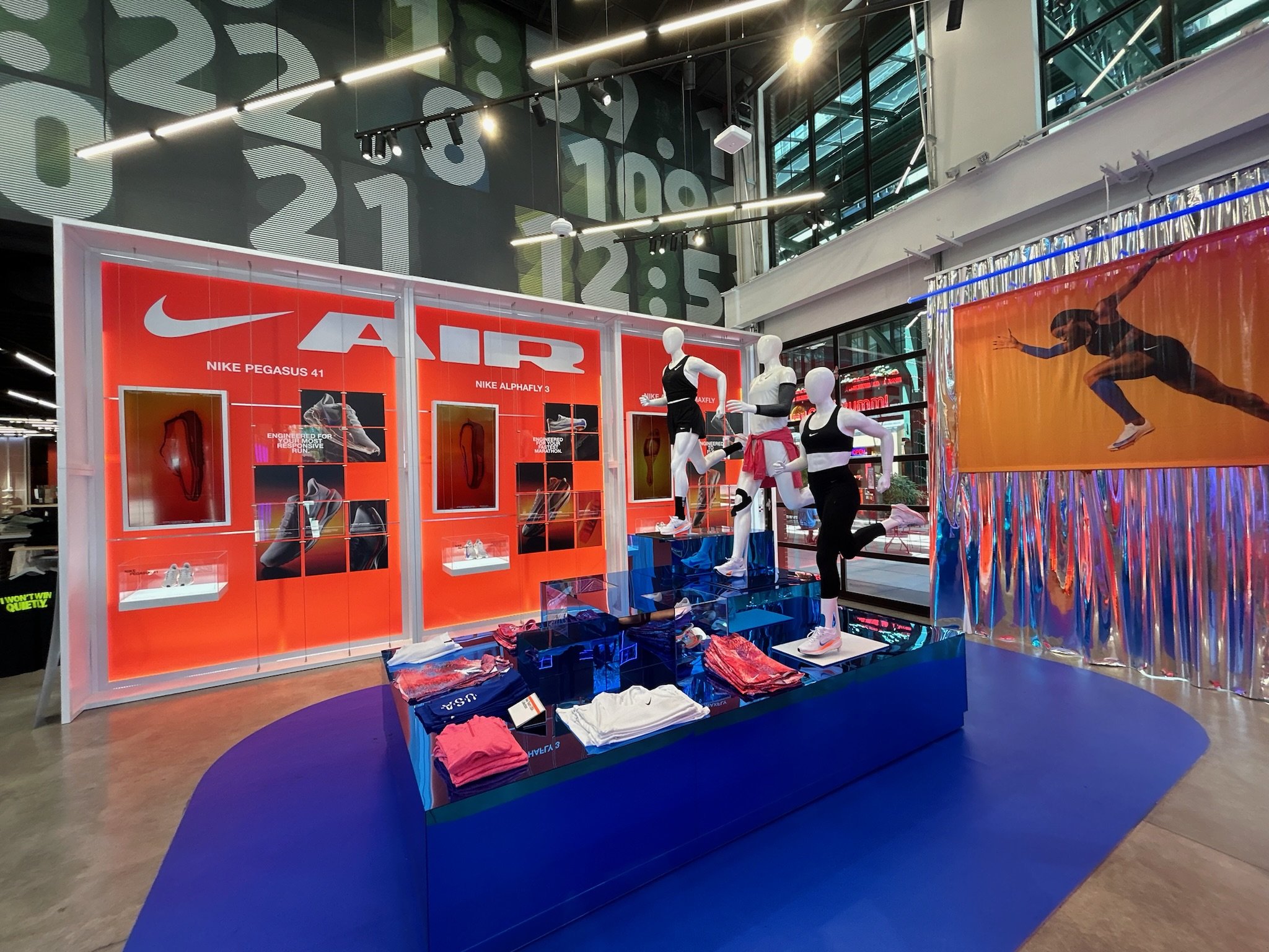 Nike store display featuring mannequins dressed in athletic wear, showcasing Nike Pegasus 41 and Nike Alphafly 3 shoes, with orange and black advertisement and a colorful backdrop with a running image.