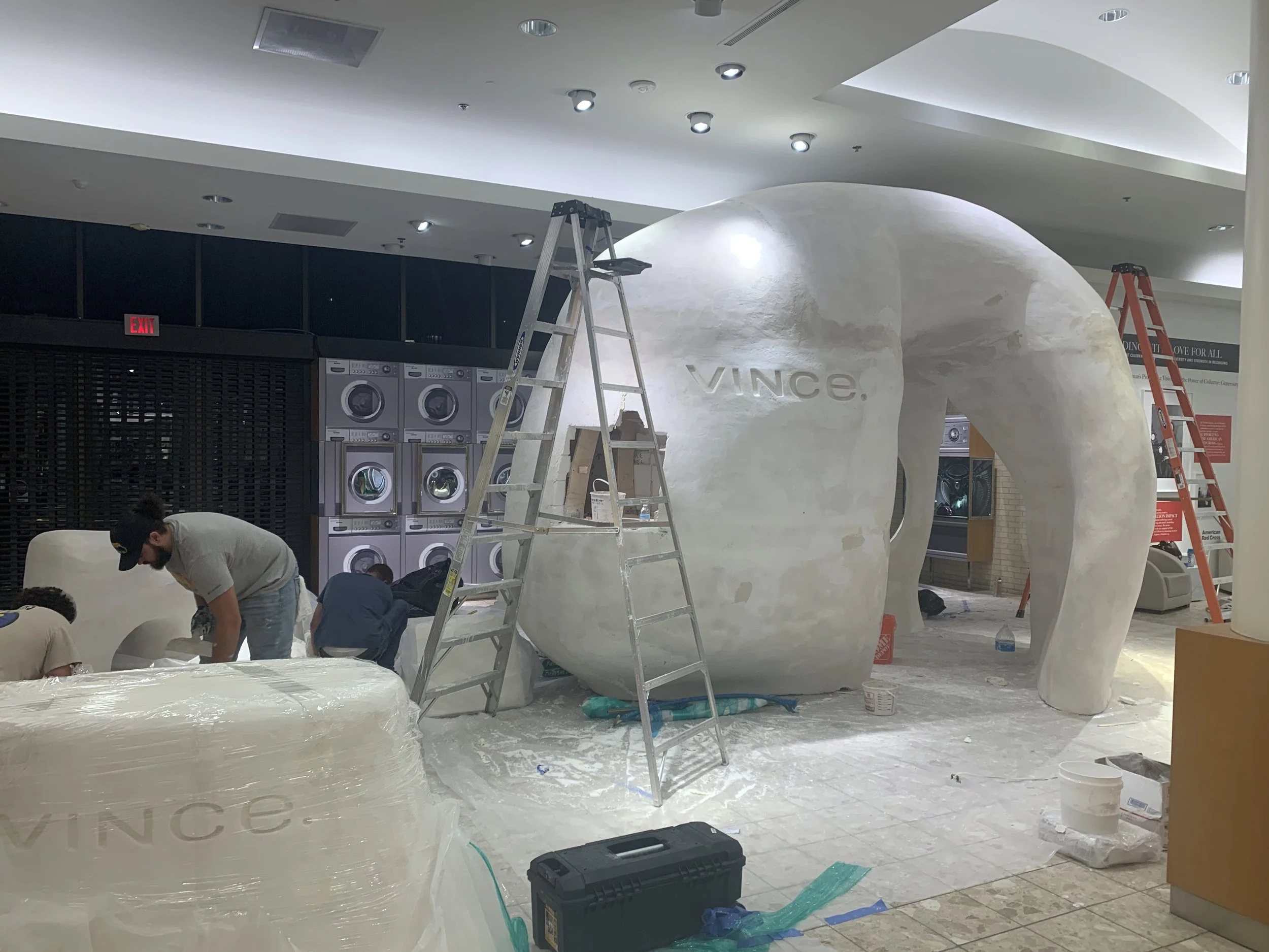 A store or mall under construction, featuring a large white sculpture resembling an elephant with the word 'Vince' carved into its side. Workers are installing or finishing the sculpture, surrounded by ladders, construction tools, and materials.