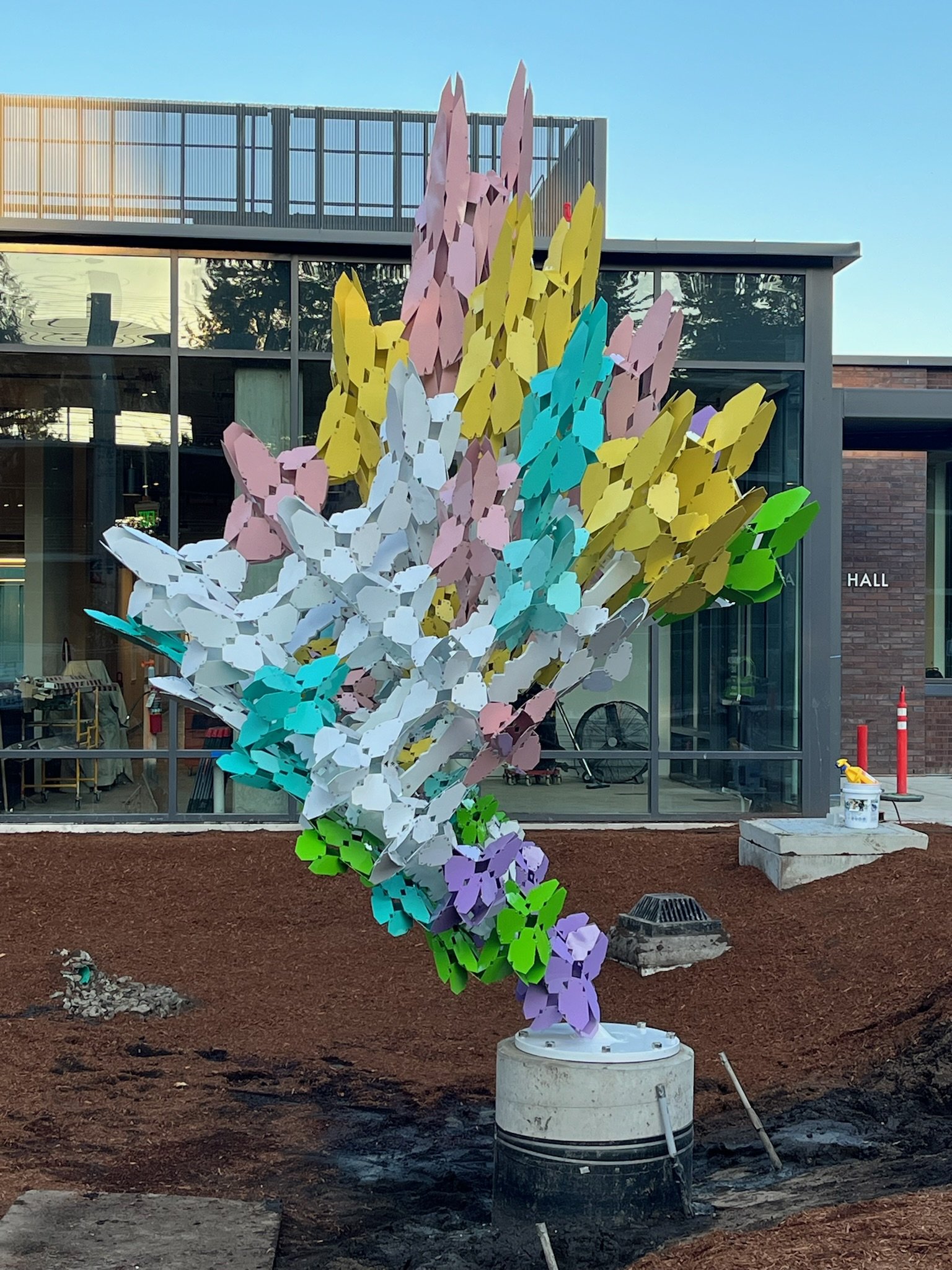 Colorful outdoor sculpture resembling a bouquet of abstract flowers in various pastel colors, placed on a construction site in front of a modern glass building.