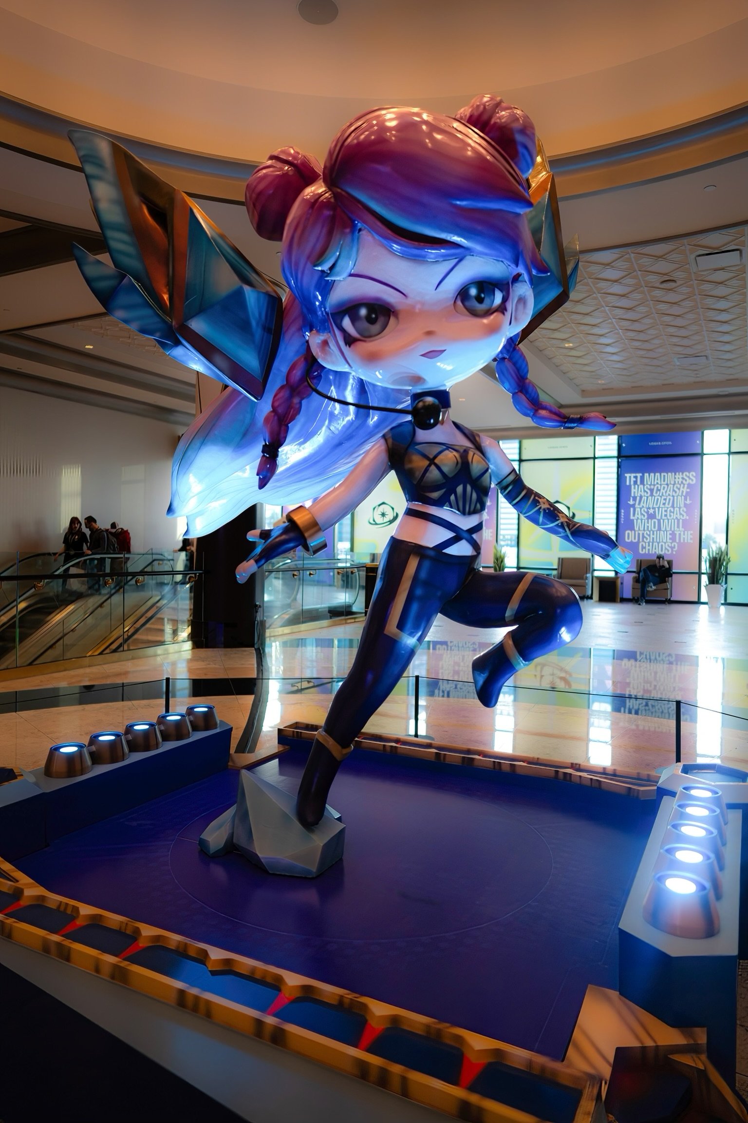 A large, stylized statue of a female fantasy character with purple hair, wearing a black and gold outfit, standing on one leg with one arm extended, in a spacious indoor setting with bright lighting and some people in the background.
