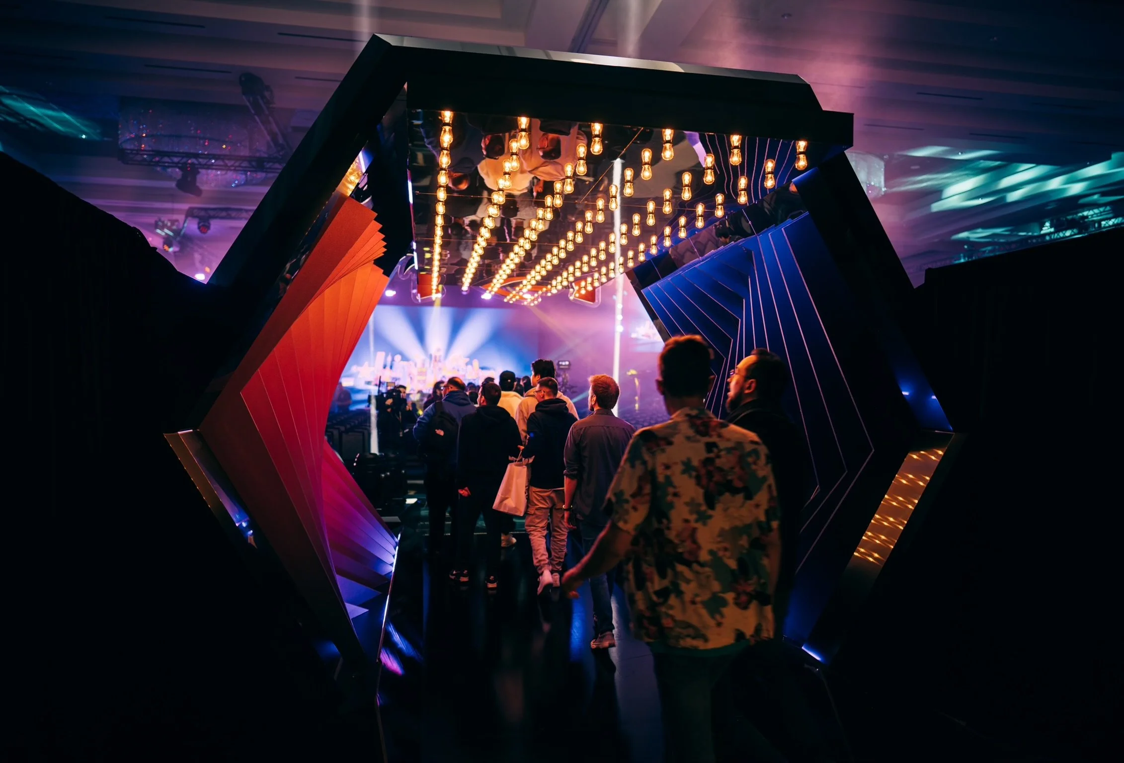 People entering a venue with decorative lighting and stage with bright lights and visual effects in the background.
