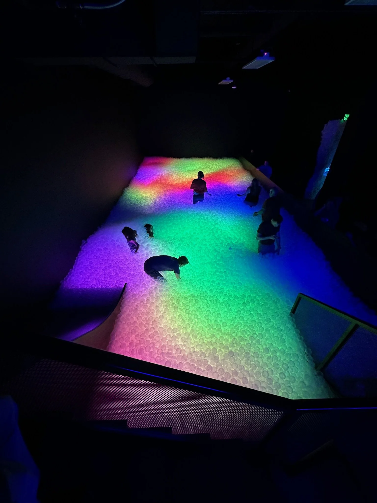 People walking on an interactive, illuminated, ball pit with rainbow-colored lighting in a dark room.