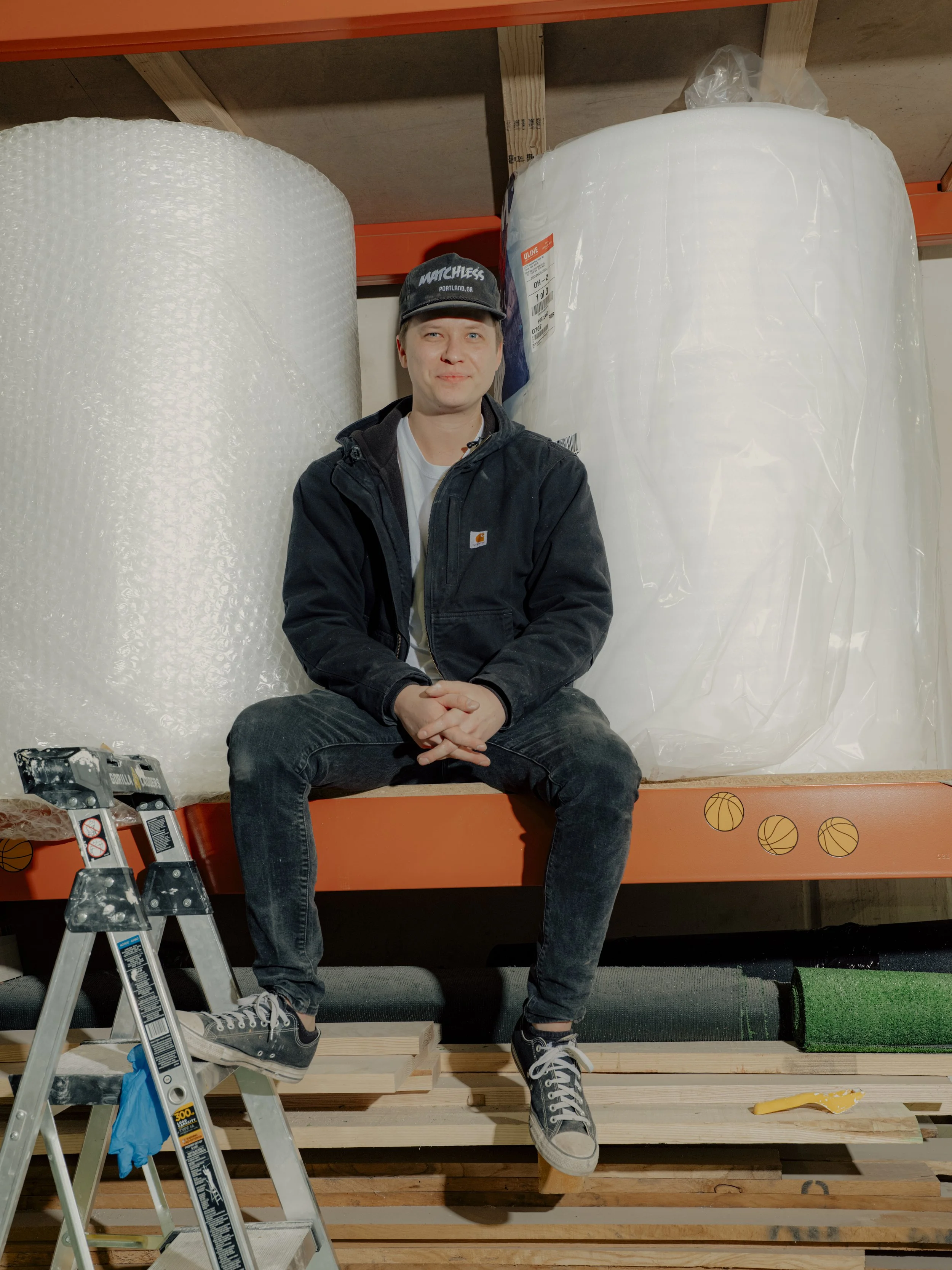A man sitting on a wooden pallet shelf in a warehouse with large rolls of bubble wrap and plastic packaging around him.
