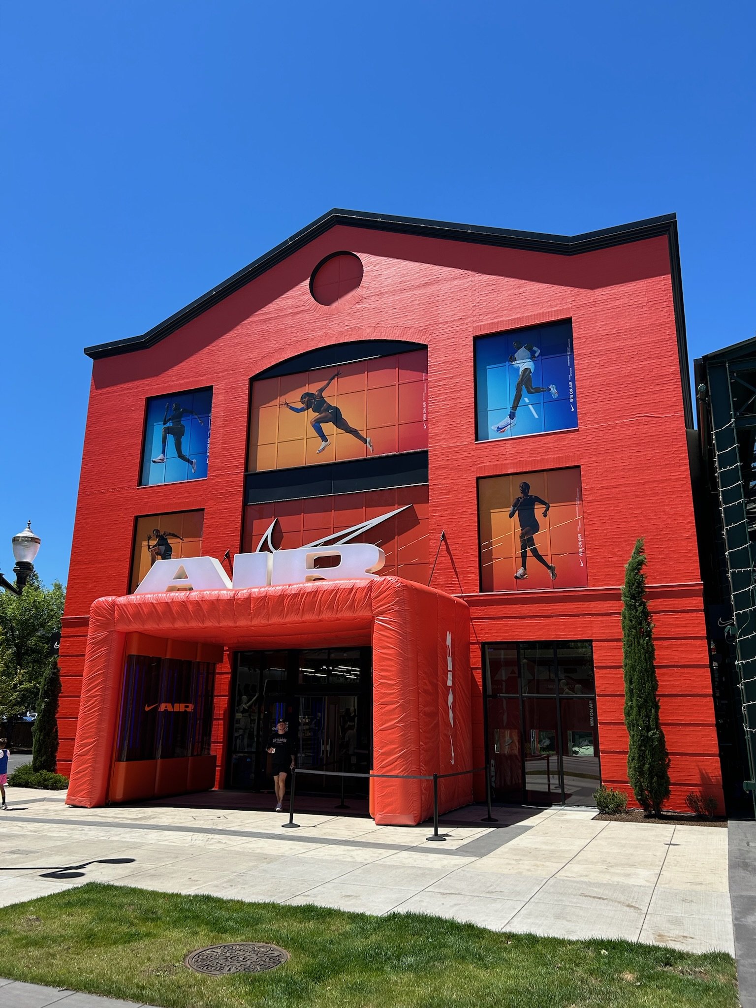Red brick Nike store with large Nike logo and images of athletes on the windows, featuring an inflatable entrance and a person walking into the store on a sunny day.