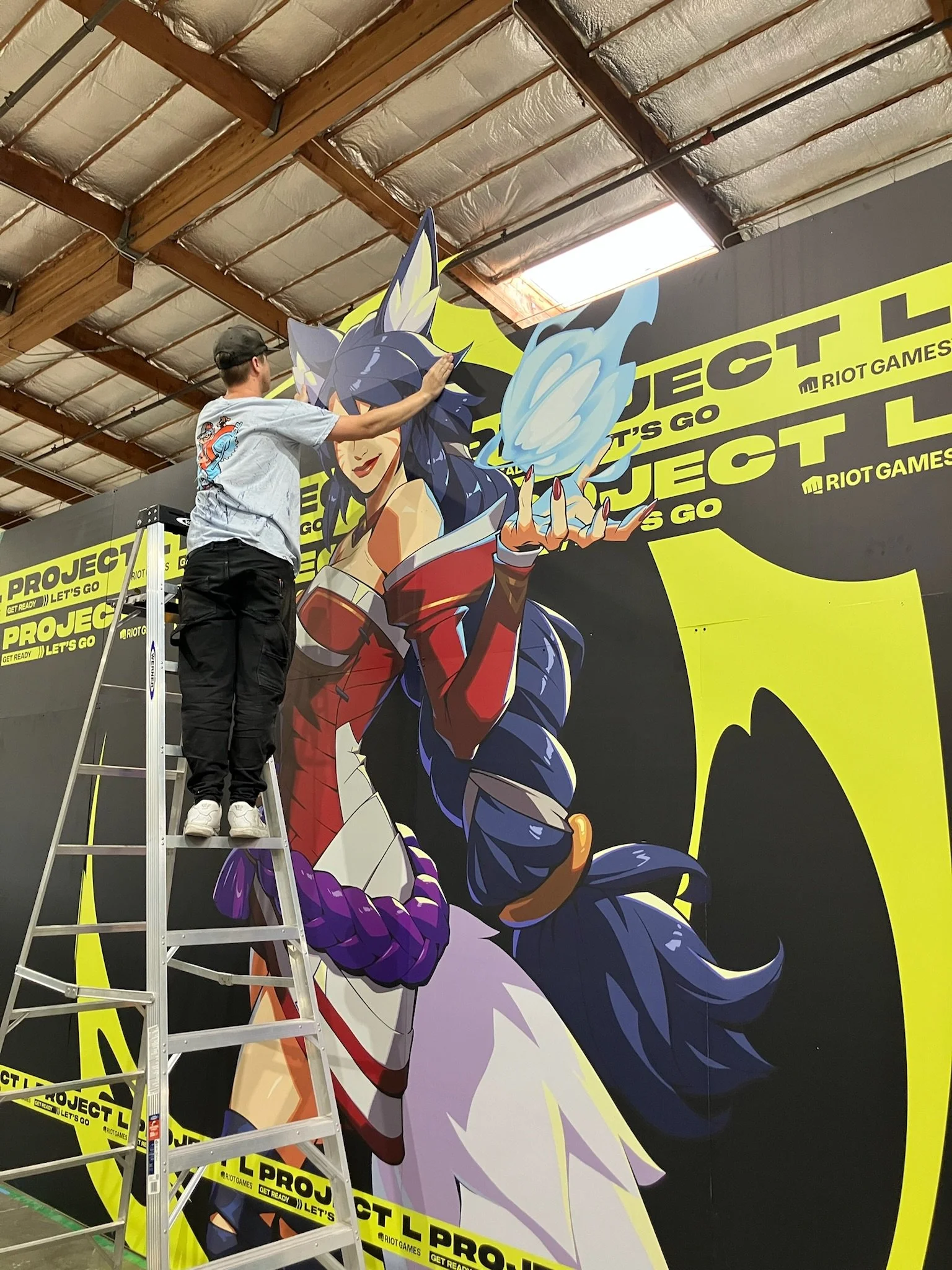A person on a ladder working on a large colorful mural of a female character from a video game, with a background of black and yellow riot games project wall.