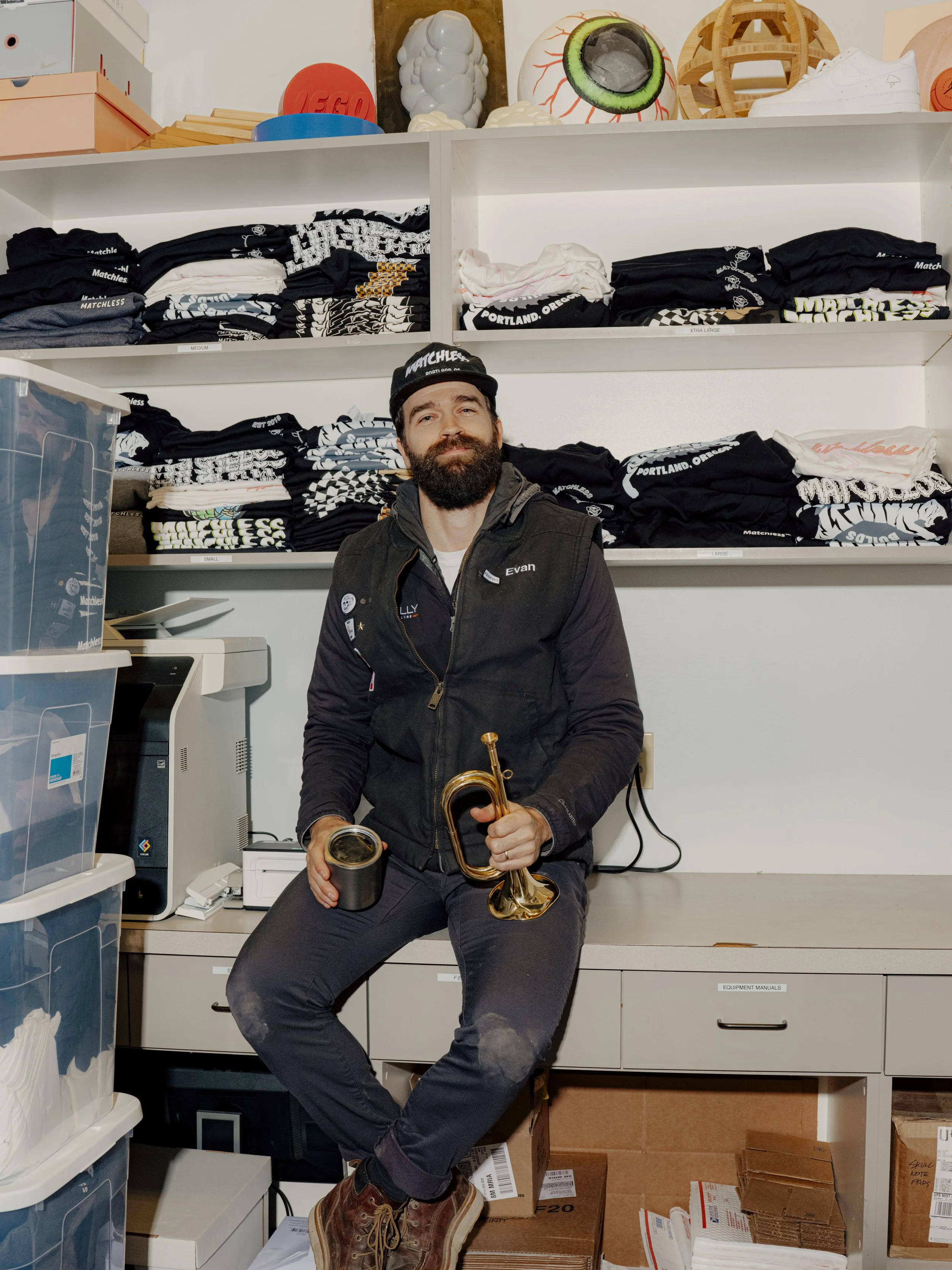 A man with a beard, wearing a black vest and black pants, sitting on a counter in a storage room. He is holding a brass instrument and a black container, smiling at the camera. Behind him are shelves with folded black and white clothing, and above are various arts and decor items.