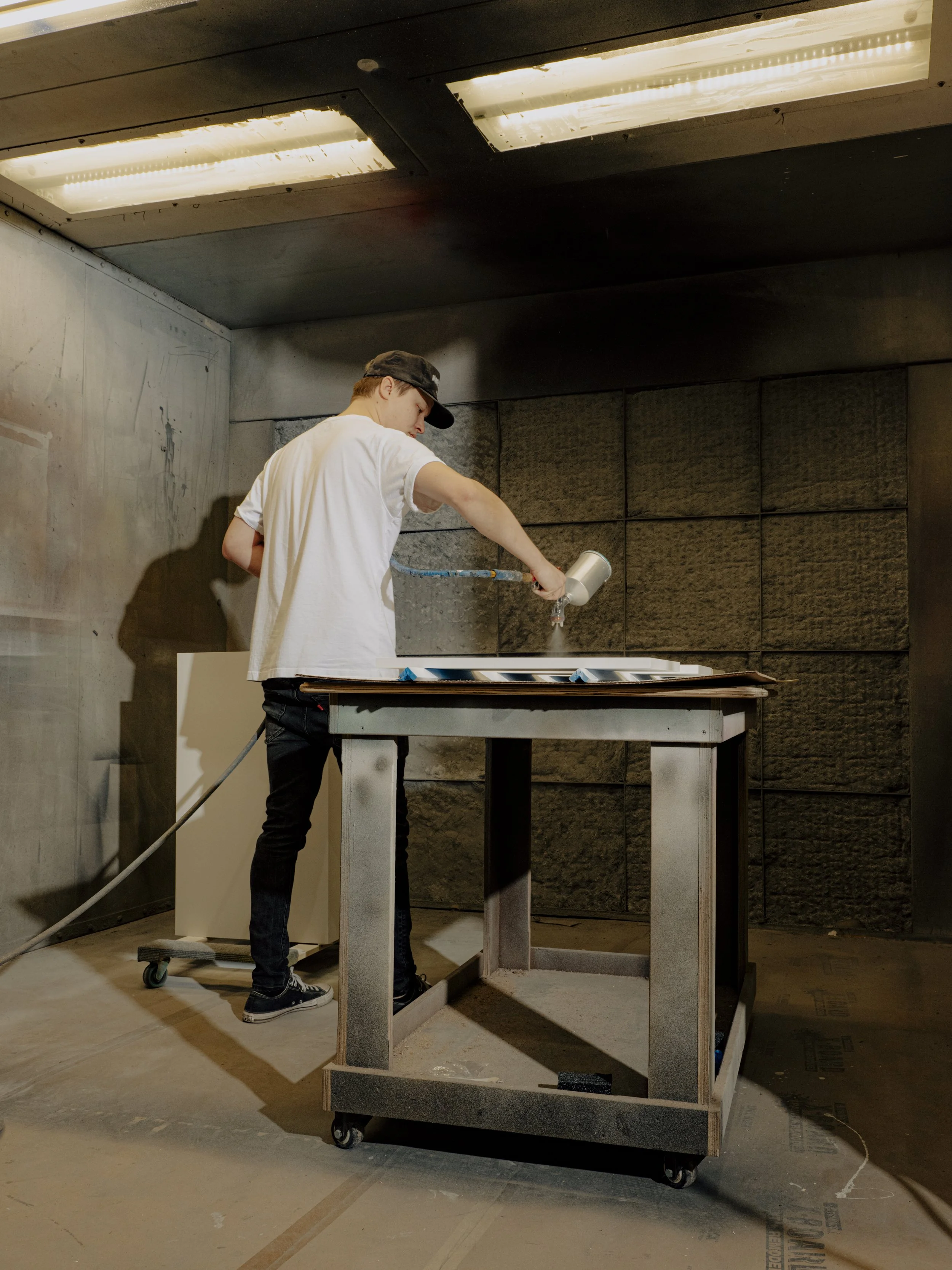 A person wearing a white T-shirt, black pants, black shoes, and a black cap is painting a wall with a spray gun in a room with dark walls and ceiling lighting.