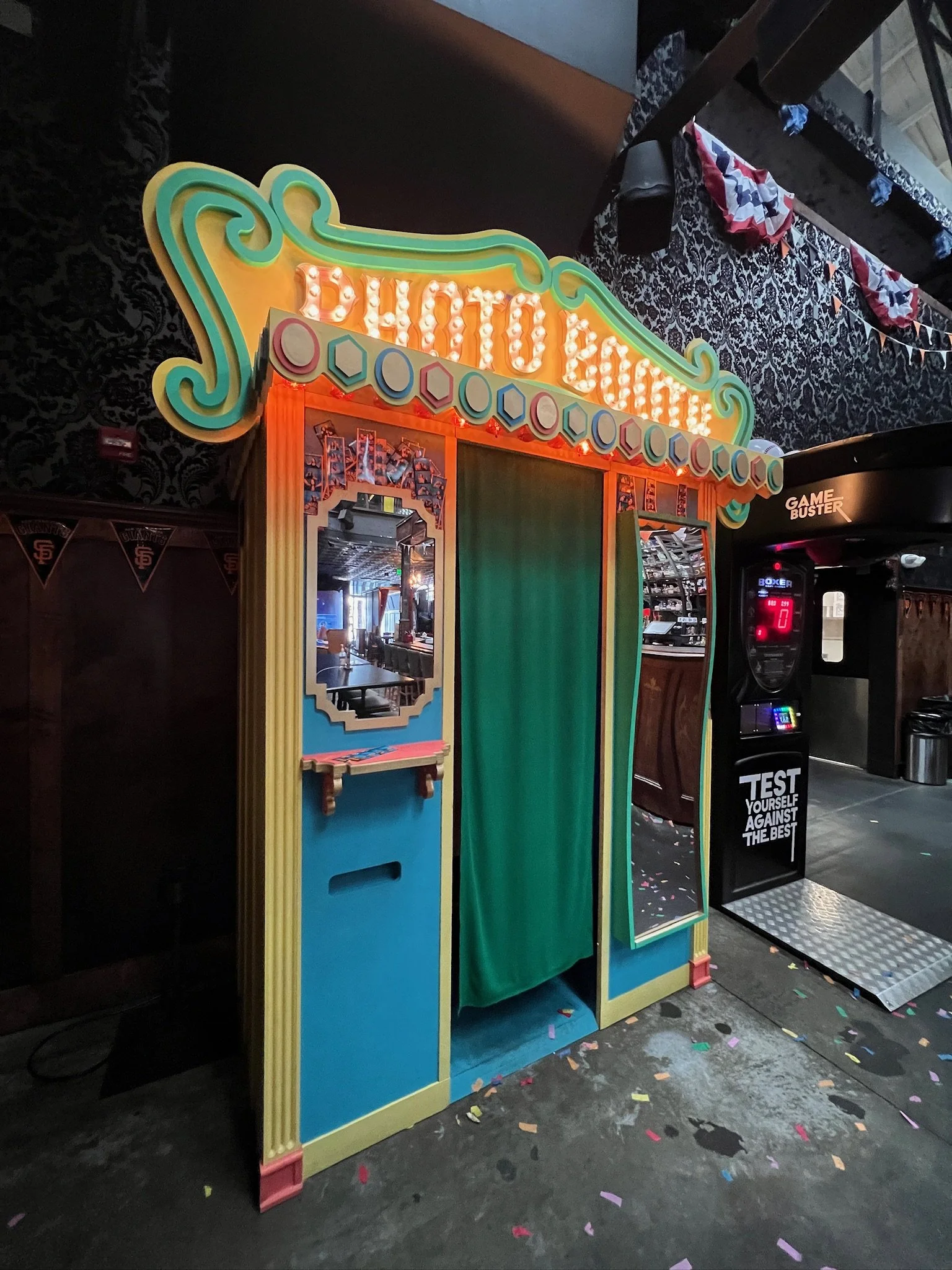 Colorful photo booth with a marquee sign that reads 'PHOTO BOOTH'. It has a green curtain for the photo area, a mirror on one side, and a small ledge below the mirror. The booth is situated on a floor with colorful confetti, next to a game arcade mac
