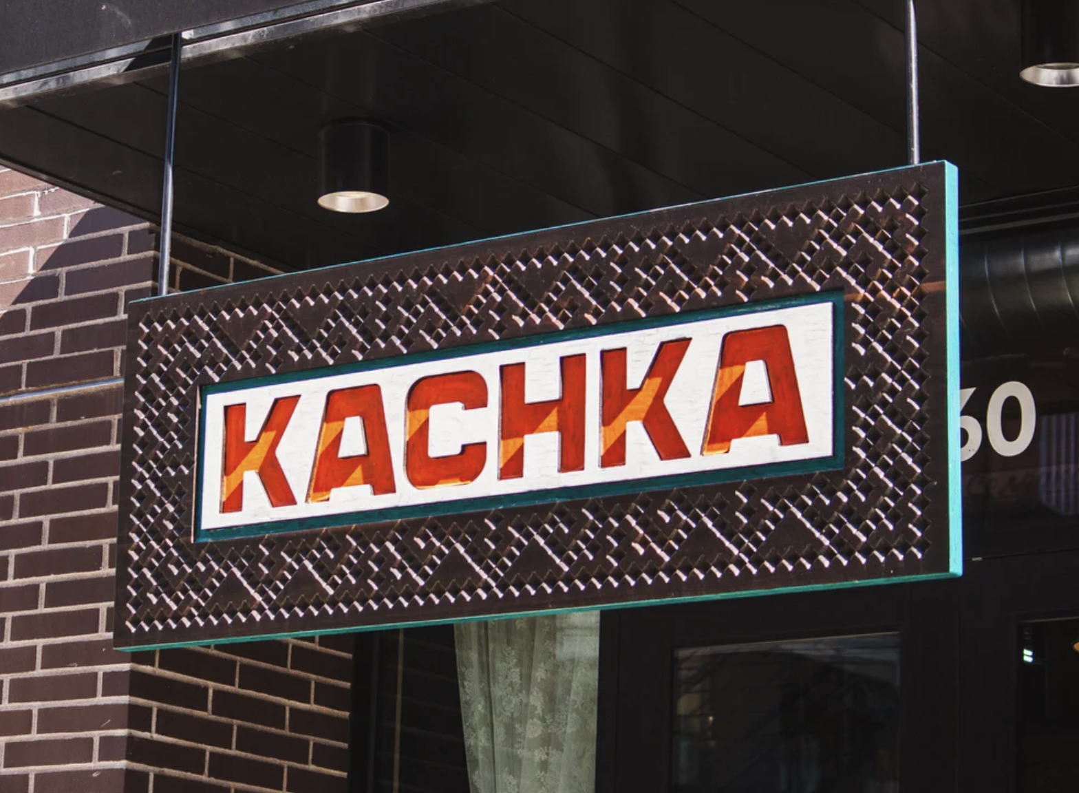 A sign with the word 'KACHKA' in red block letters on a white background, mounted on a black decorative lattice outside a building with a brick wall and a large window.