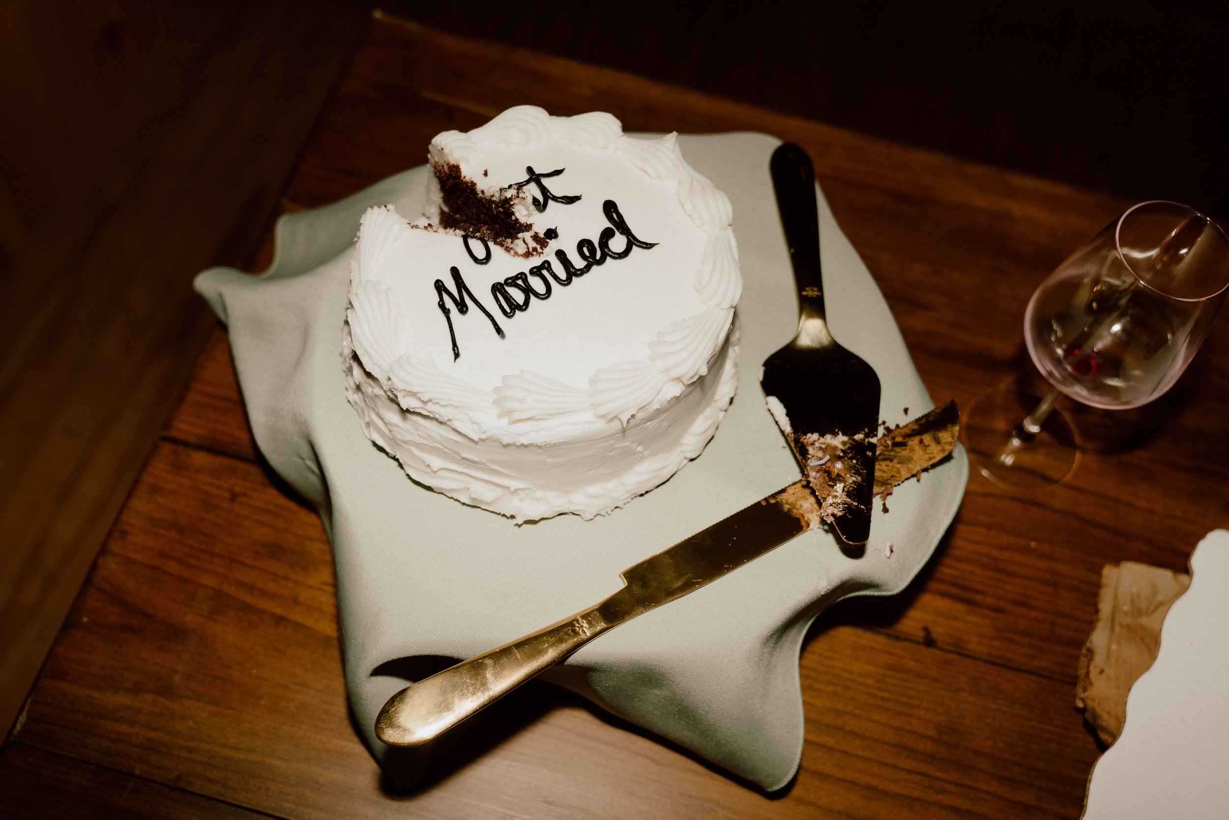 A partially eaten wedding cake with white frosting, decorated with piped borders, and the words "Just Married" written on top in chocolate. The cake is on a silver cake board, with a slice and a cake knife nearby, and a wine glass with some red wine beside it, all on a wooden table.