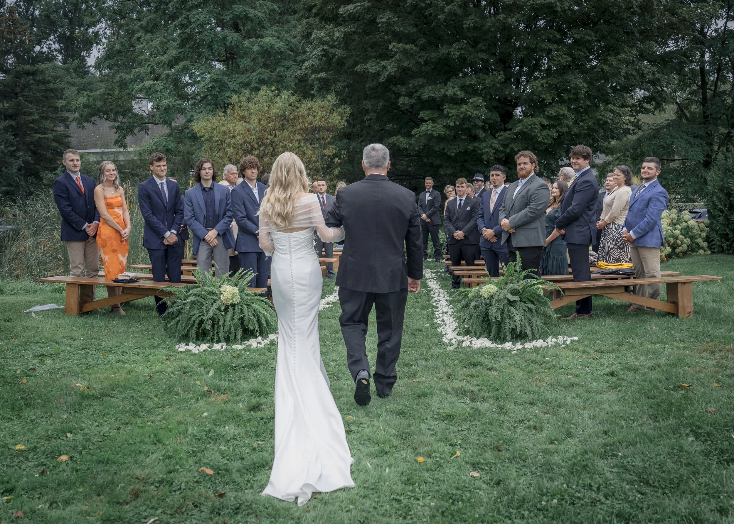 A couple is walking down the aisle at an outdoor wedding ceremony, with guests seated on either side of the aisle.