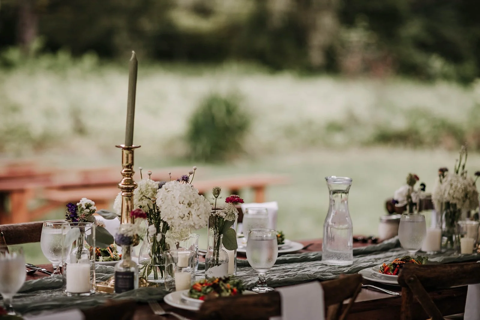 A decorated outdoor dining table with floral centerpieces, bottles, glasses of water, a candle, a candlestick, plates with food, and a grey table runner, set in a natural garden setting.